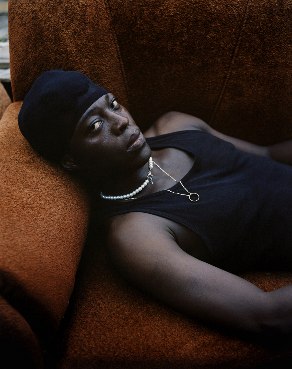 a ghanian man in a black tank top and jewelry lounging on an orange sofa