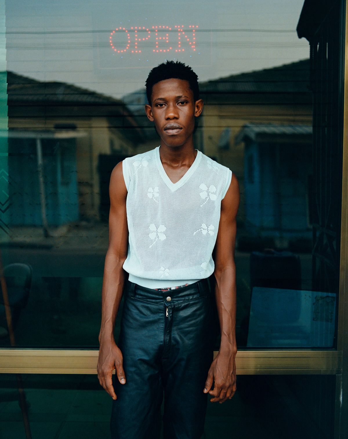 ghanian youth in a white floral vests and leather pants posing for a portrait in front of an open sign