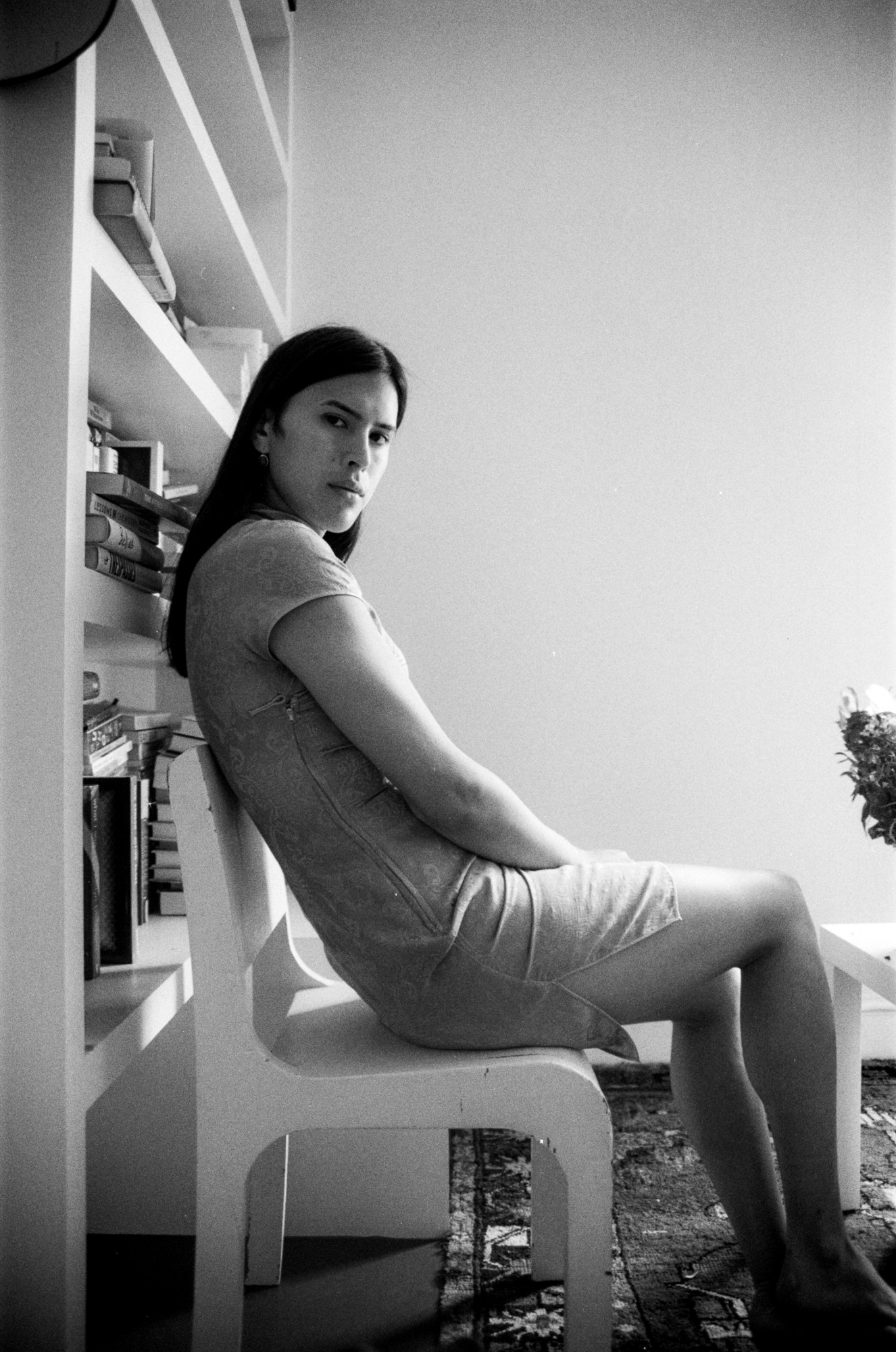 a black and white portrait of the author nicola dinan in her living room wearing a silk dress. she's sat on a white chair and is looking over her shoulder towards the camera.