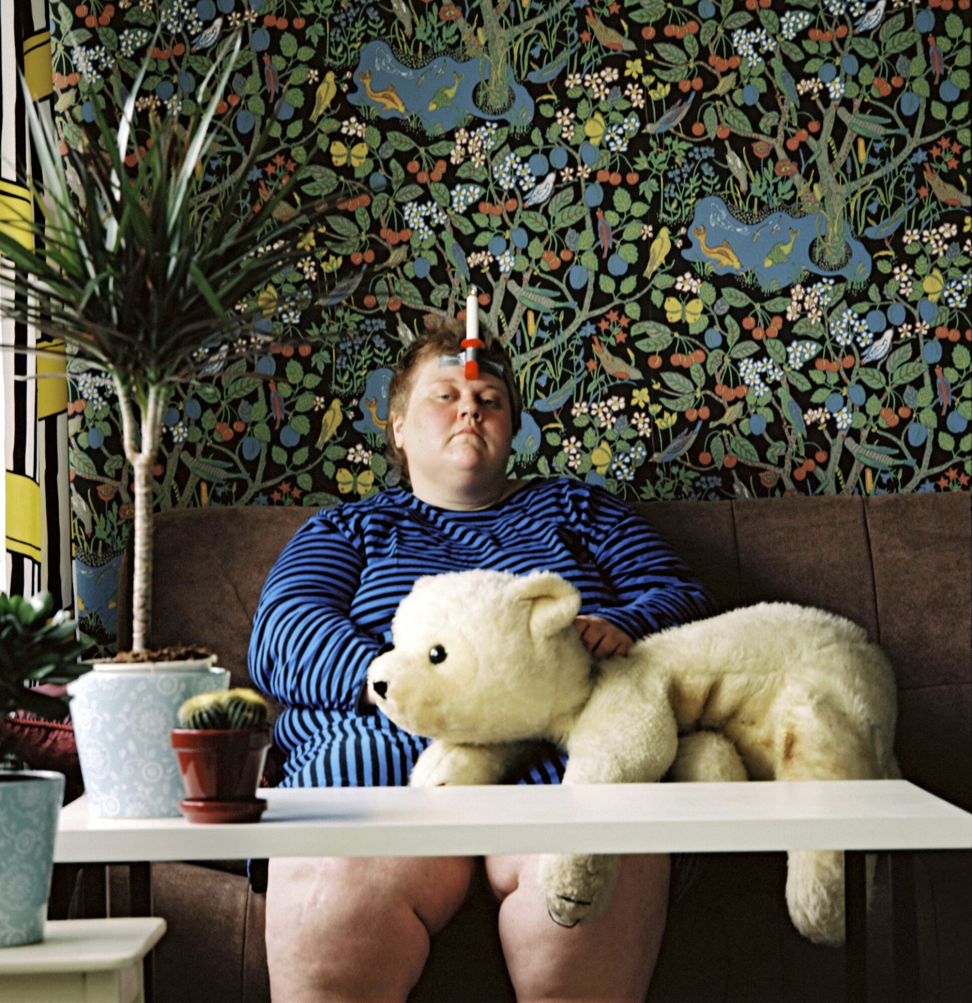 A woman holding a large white teddy bear with a candle taped to her forehead