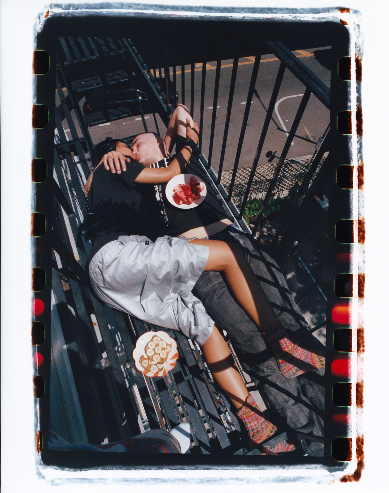 a couple embrace laying down on a NYC fire escape. breakfast plates of toast and fruit lay by their side