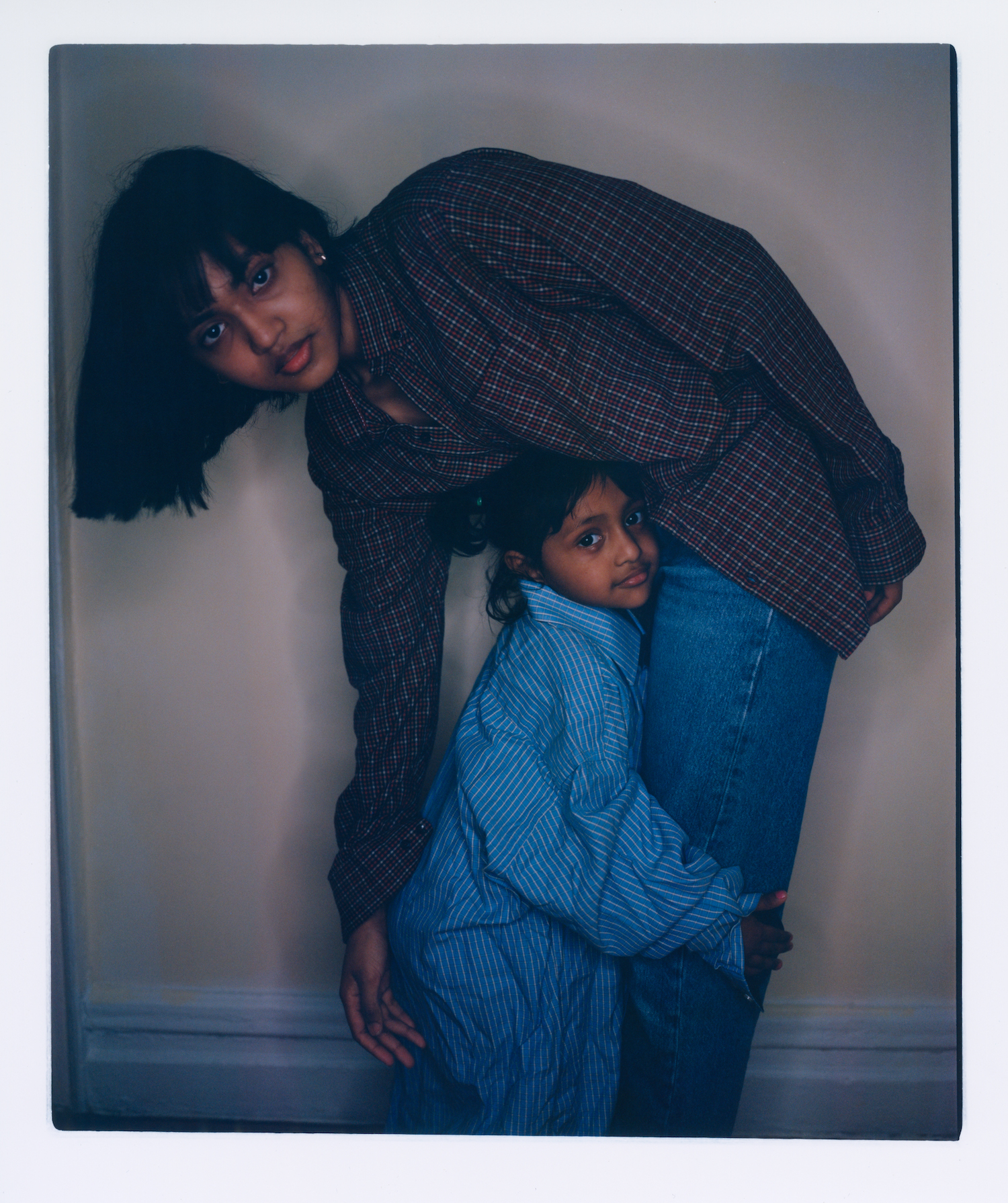 a young bangladeshi woman in a checked shirt and jeans leans over to her 3 year old sister, dressed in an oversized shirt, who is hugging her legs