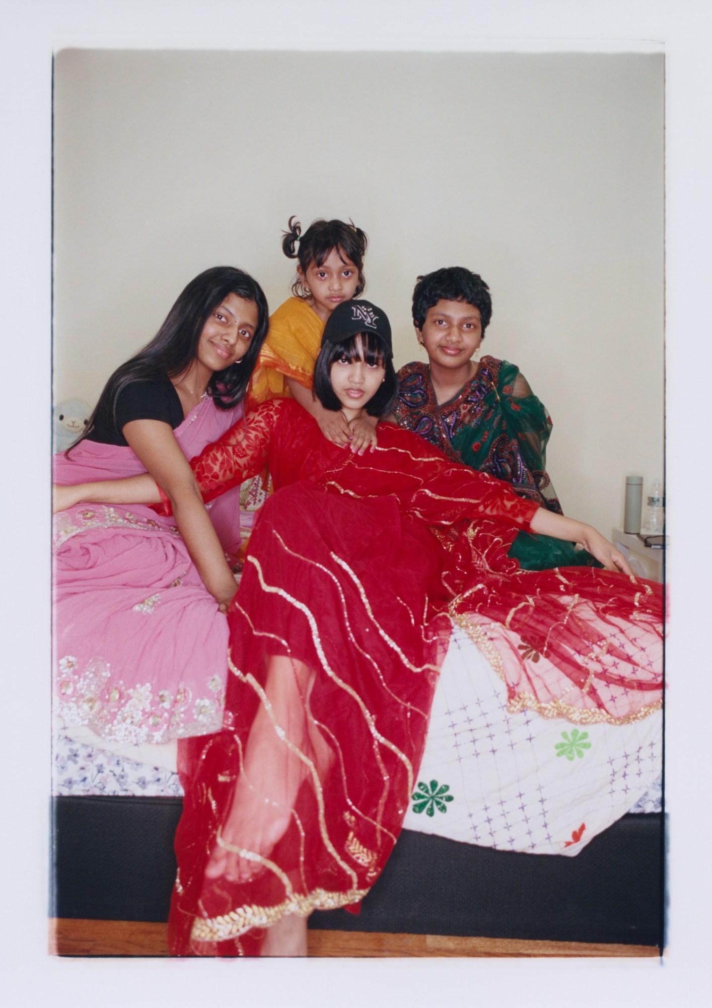 four bangladeshi sisters between the ages of 3 and 21 sit together on a bed wearing colourful formal dress, the eldest sister in the centre also wears a NY baseball cap