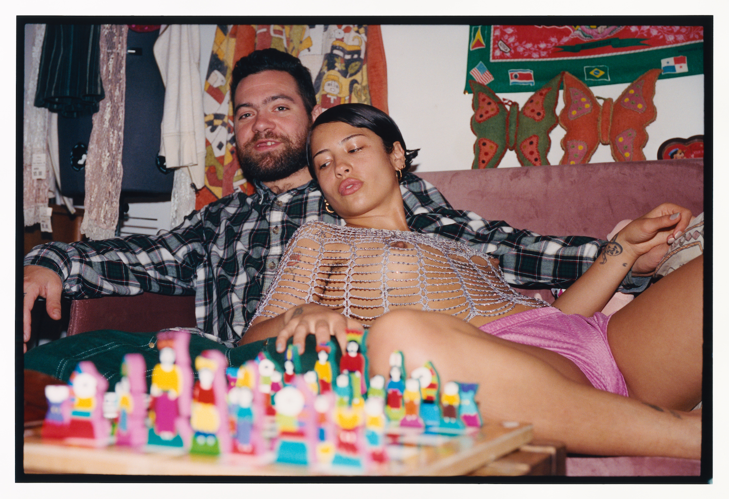 a woman in a silver cobweb-style top and pink briefs reclines on her male partner on a sofa in front of a colourful chess game; he appears to be missing one front tooth