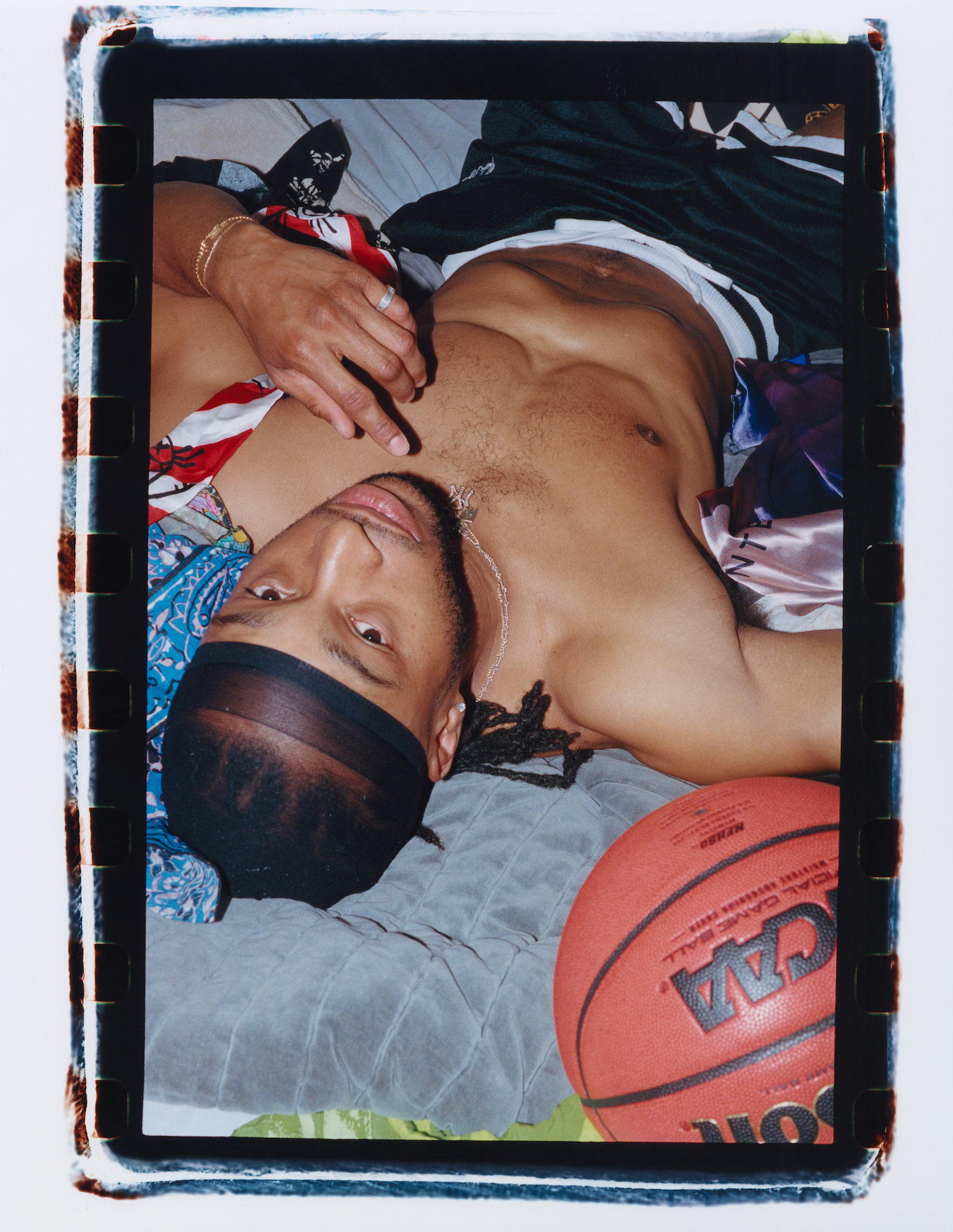 a young black man wearing a durag, a NY chain and basketball shorts lays down on his bed looking to camera