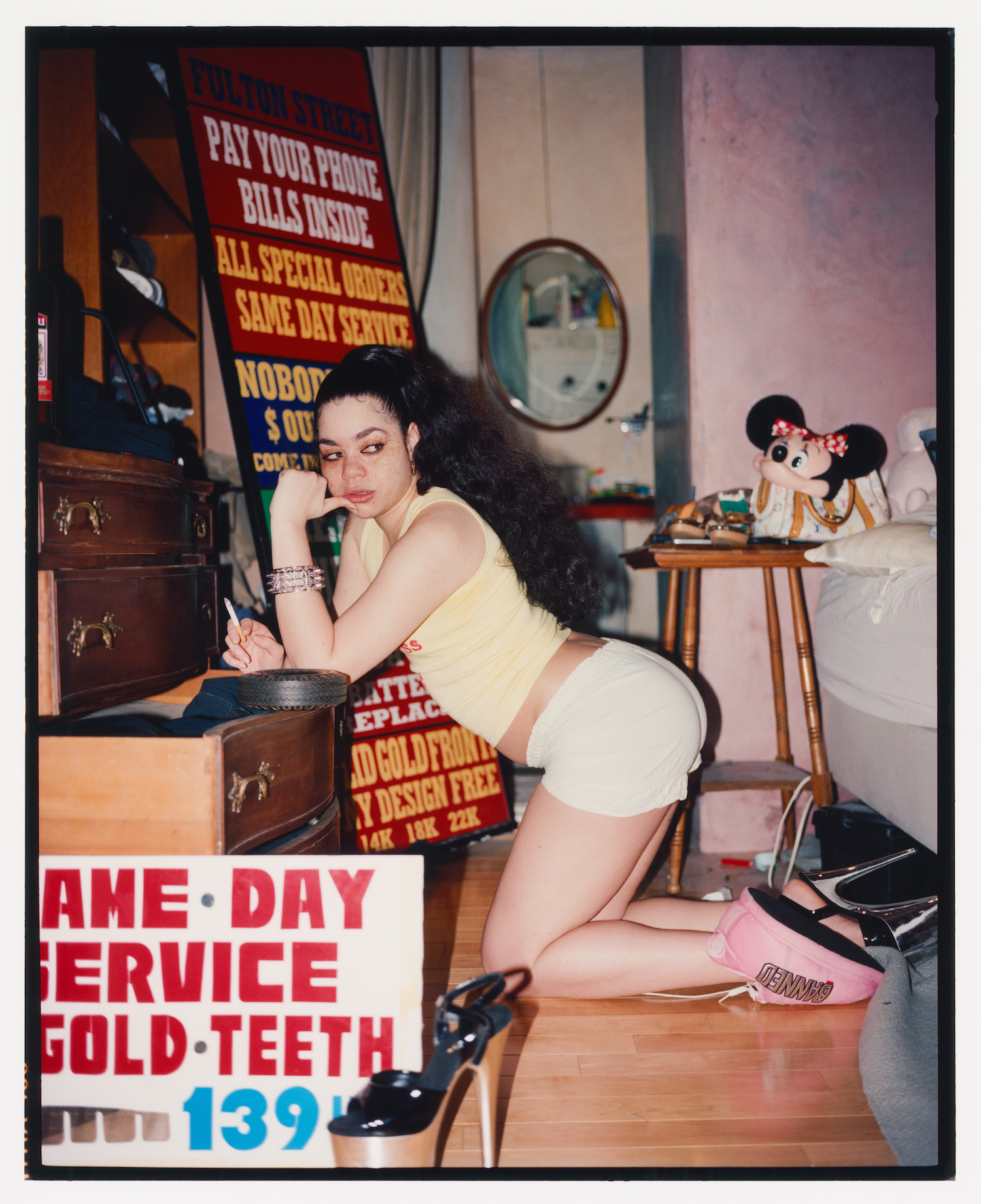 a woman with long curly hair poses smoking in a busy bedroom surrounded by vintage signs and other knick-knacks