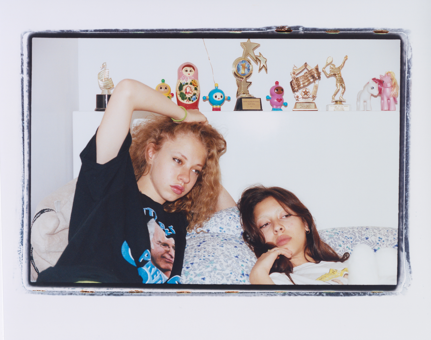 two women in oversized T-shirts pose on their bed in front of a shelf of trophies and childhood toys