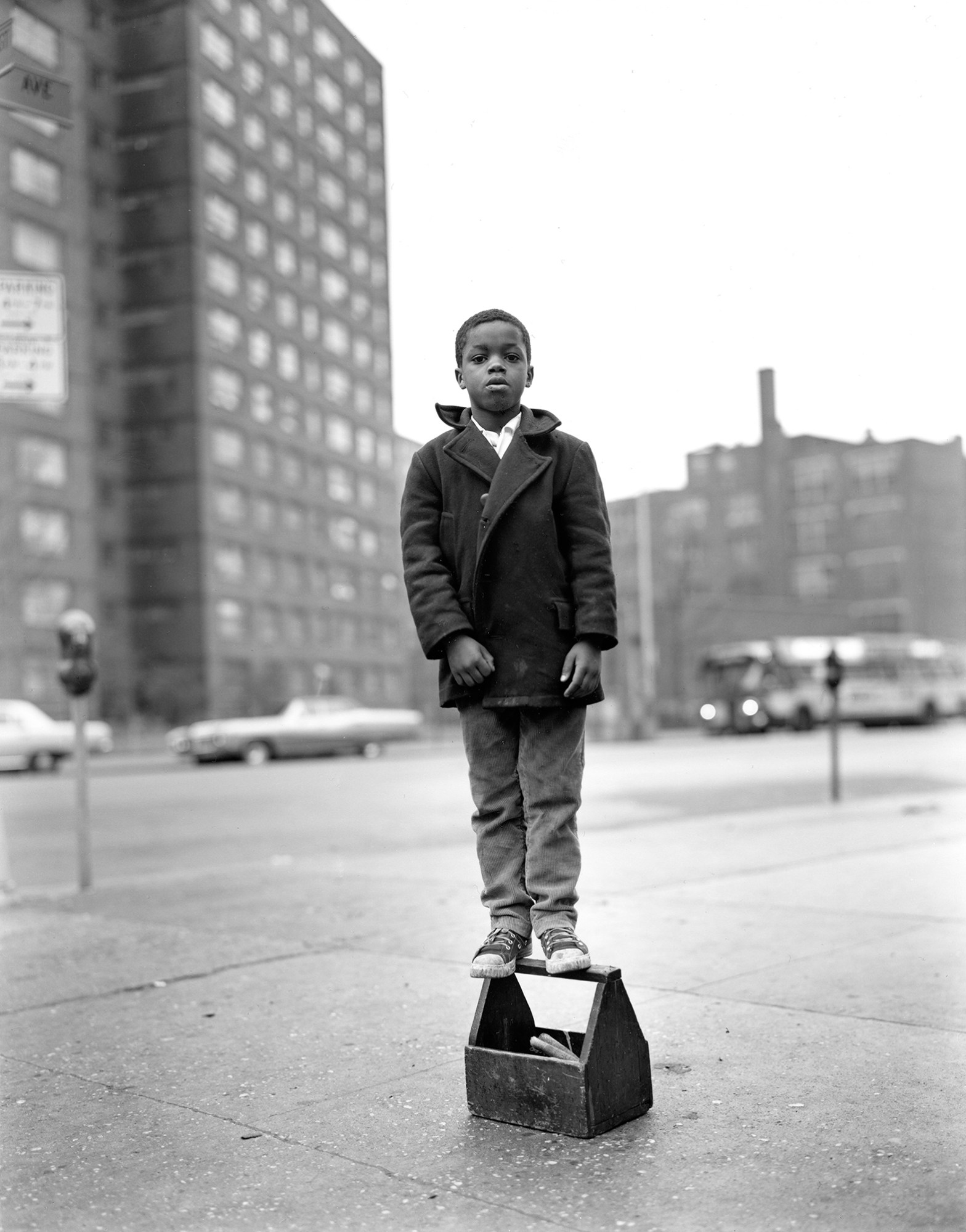 a young Black boy stands on a wooden toolbox in an empty city street; he wears an overcoat and a pair of chuck taylors