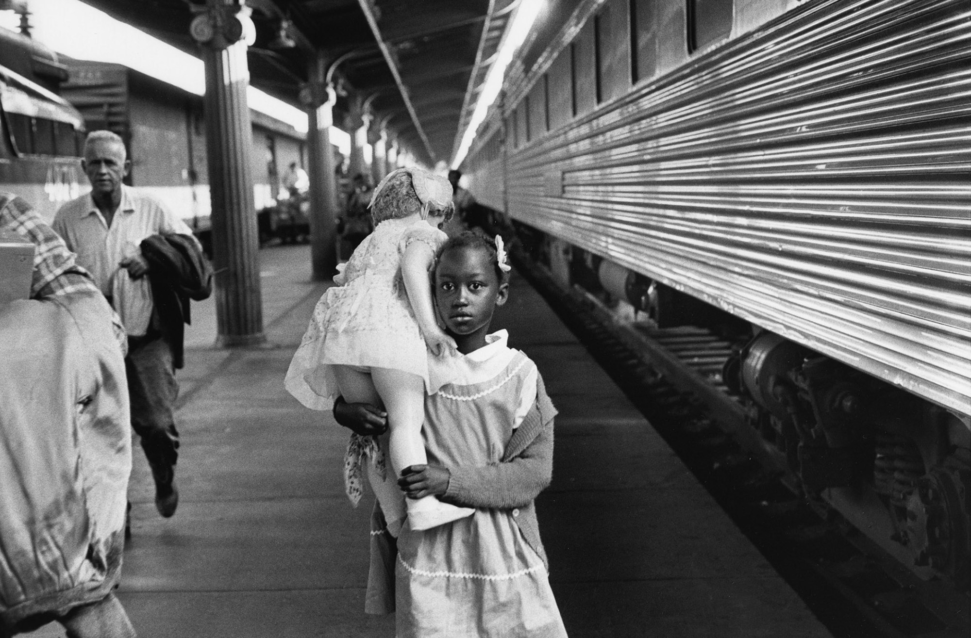 a young black girl holding a large white doll over her shoulder stands on a train platform looking to camera