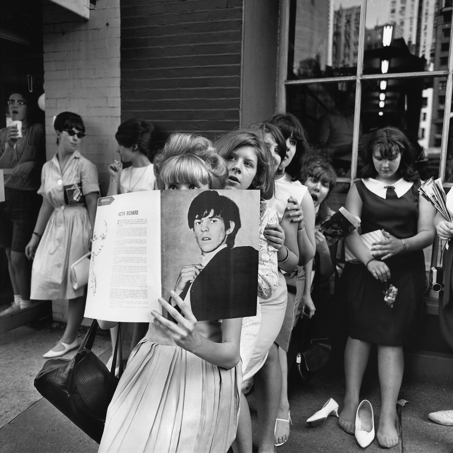 a group of young female fans stand outside queueing for an event; one young woman holds up a keith richard fanzine