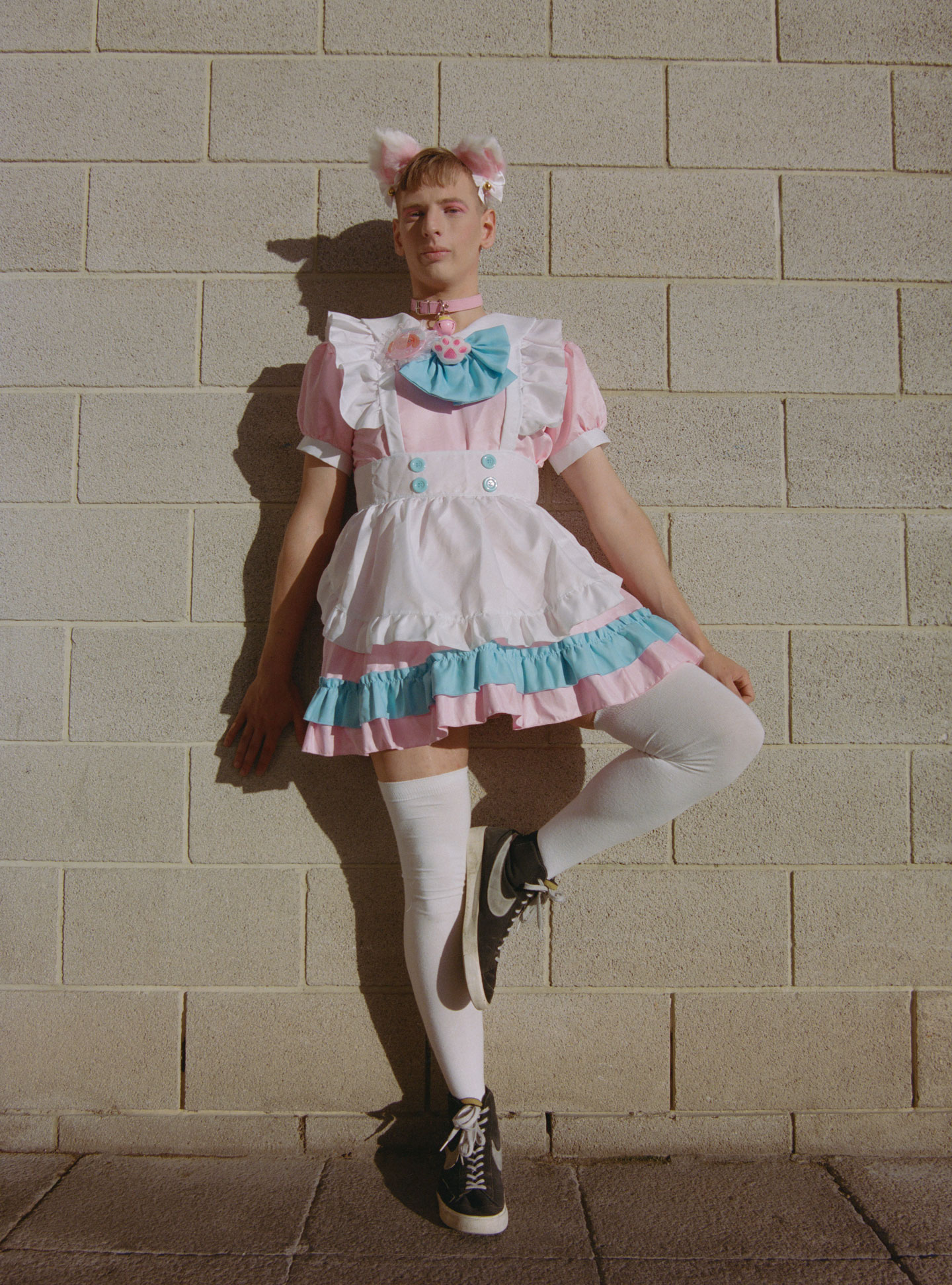 a young person with cropped blonde hair wears a cutesy maid cafe look, posing with their leg flamingoed against a wall