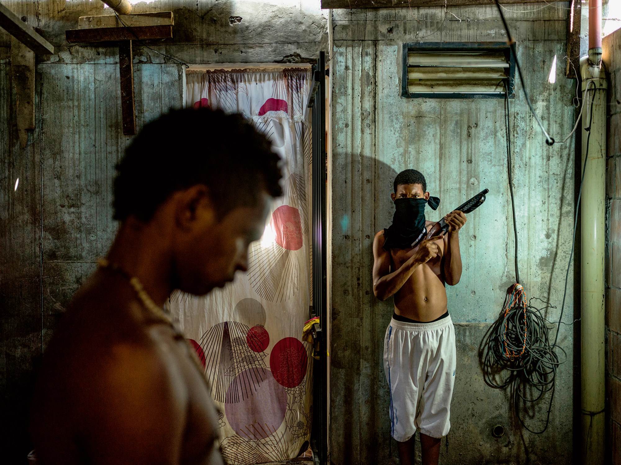 two young men stand in a room with concrete walls with hanging wires and exposed pipes; one of them stands against a wall, tshirt wrapped around his face, holding a gun