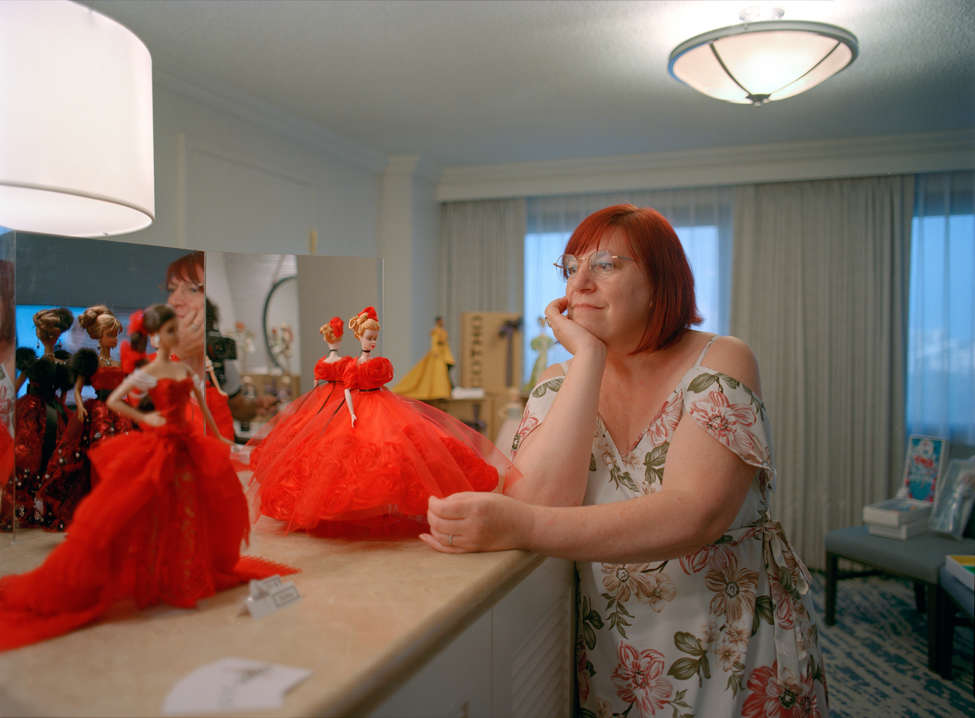 a woman in a print floral dress standing next to a collection of barbie dolls in red dresses