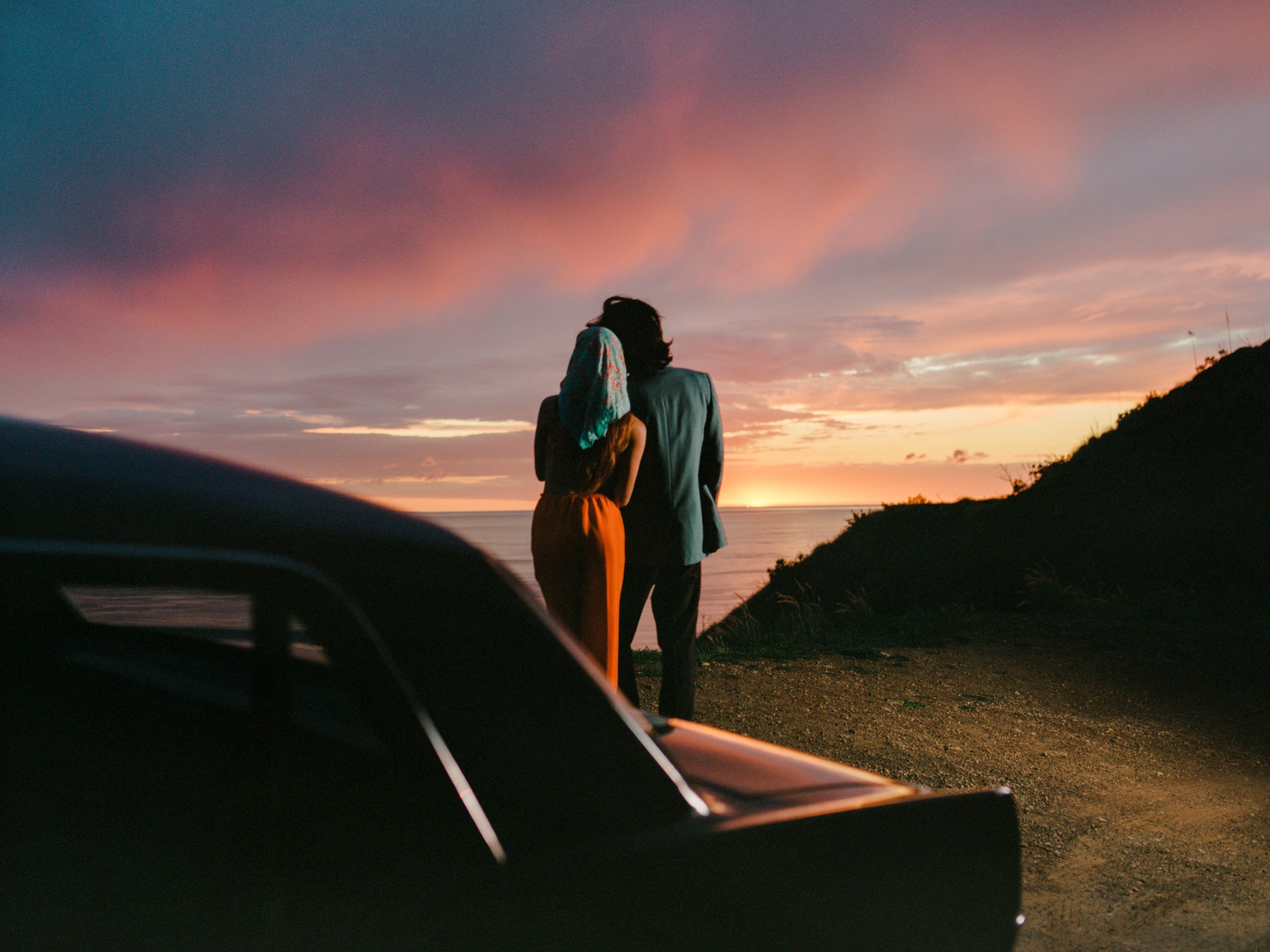 a couple staring away from the camera towards the sunset with a car in the background