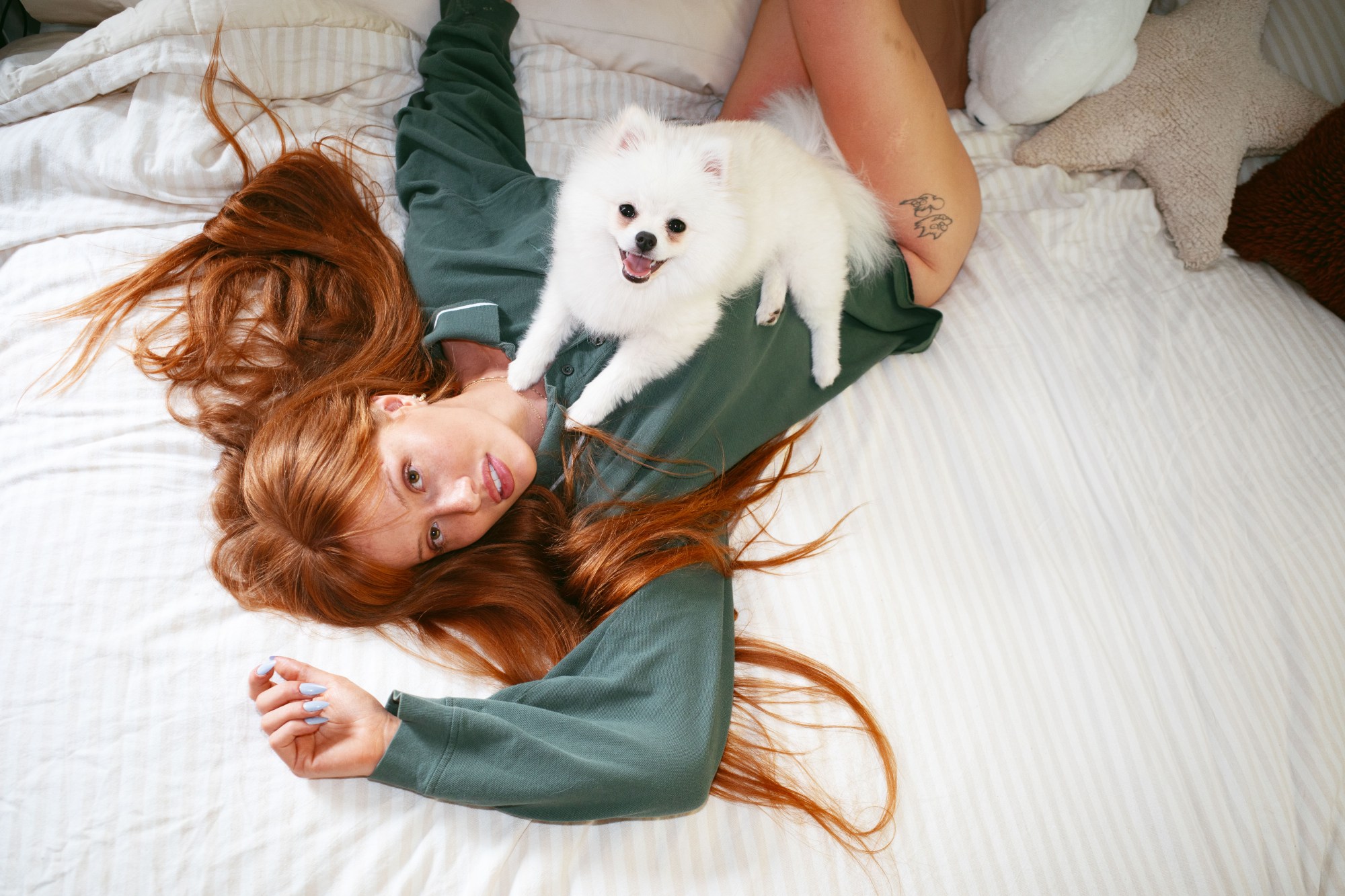 helena lying on her bed in a sweatshirt with her white pomeranian photographed by morgan maher