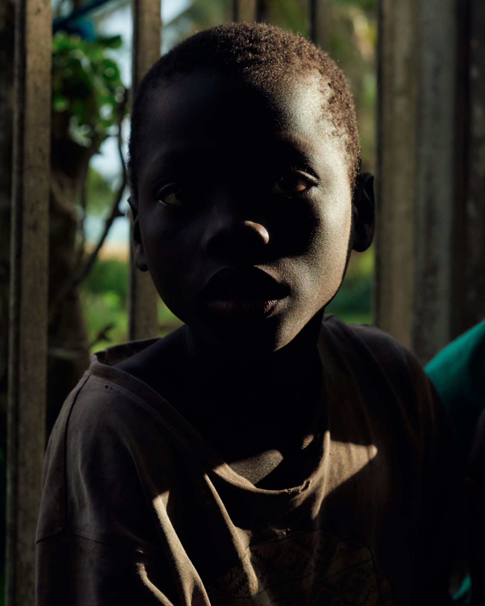 a young Black child in an oversized brown T-shirt looks to the camera, half obscured by shadow