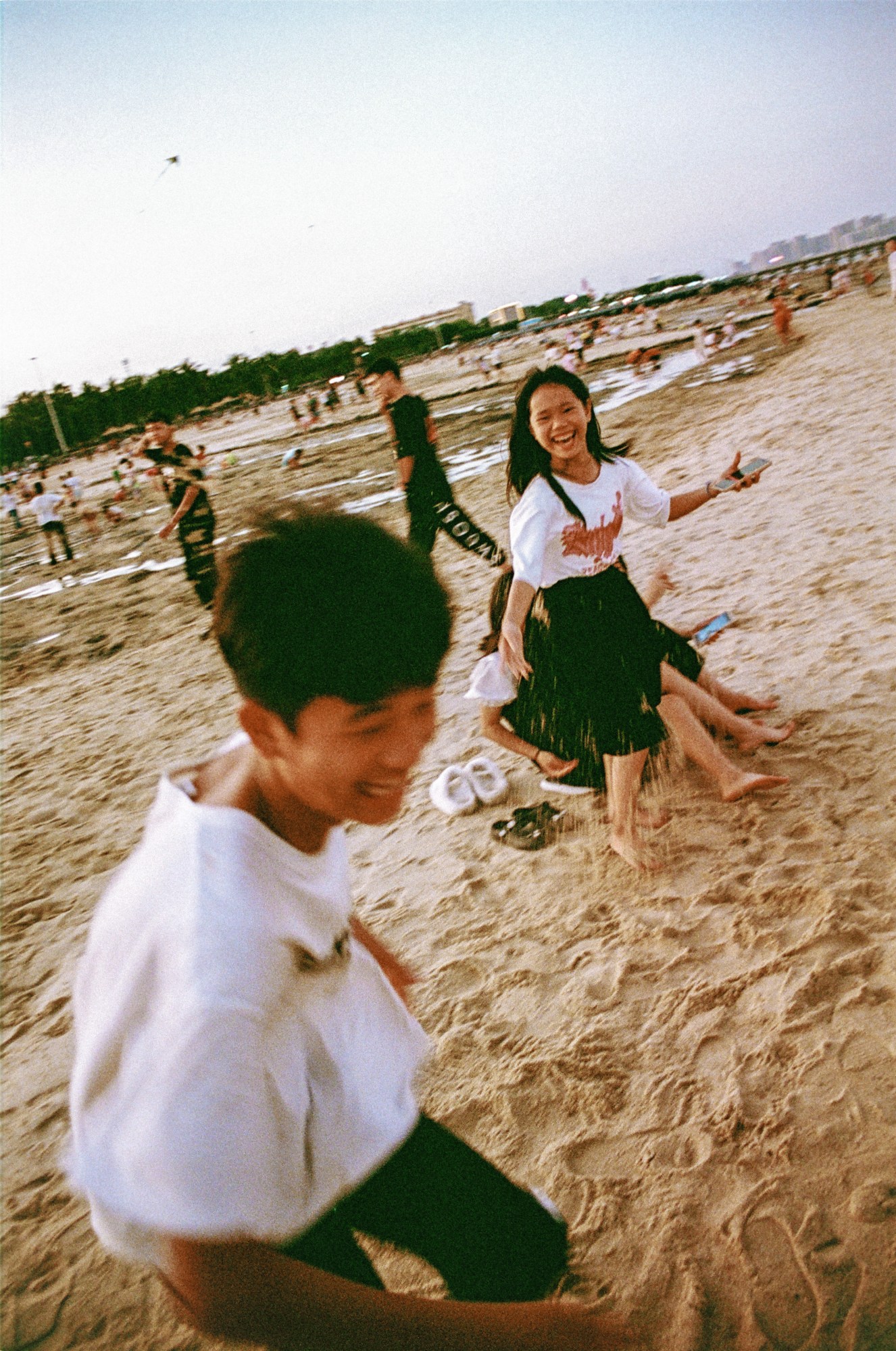 Two young, fully clothed people run together on a busy beach; they're laughing and their movement is slightly out of focus.