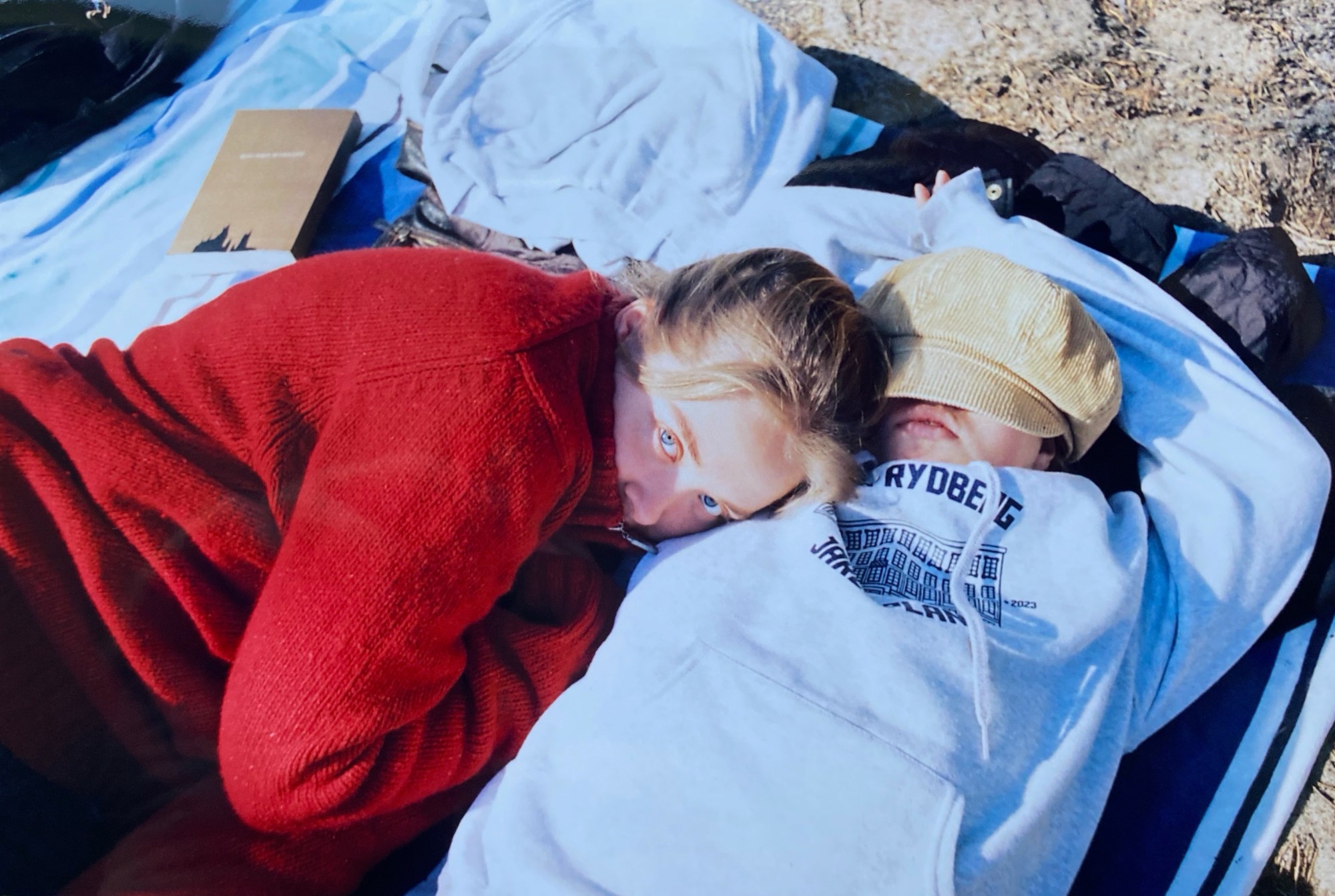 Two Swedish teens (one in a red knit sweater and the other in a hoodie and baker boy hat) lay on a beach blanket together