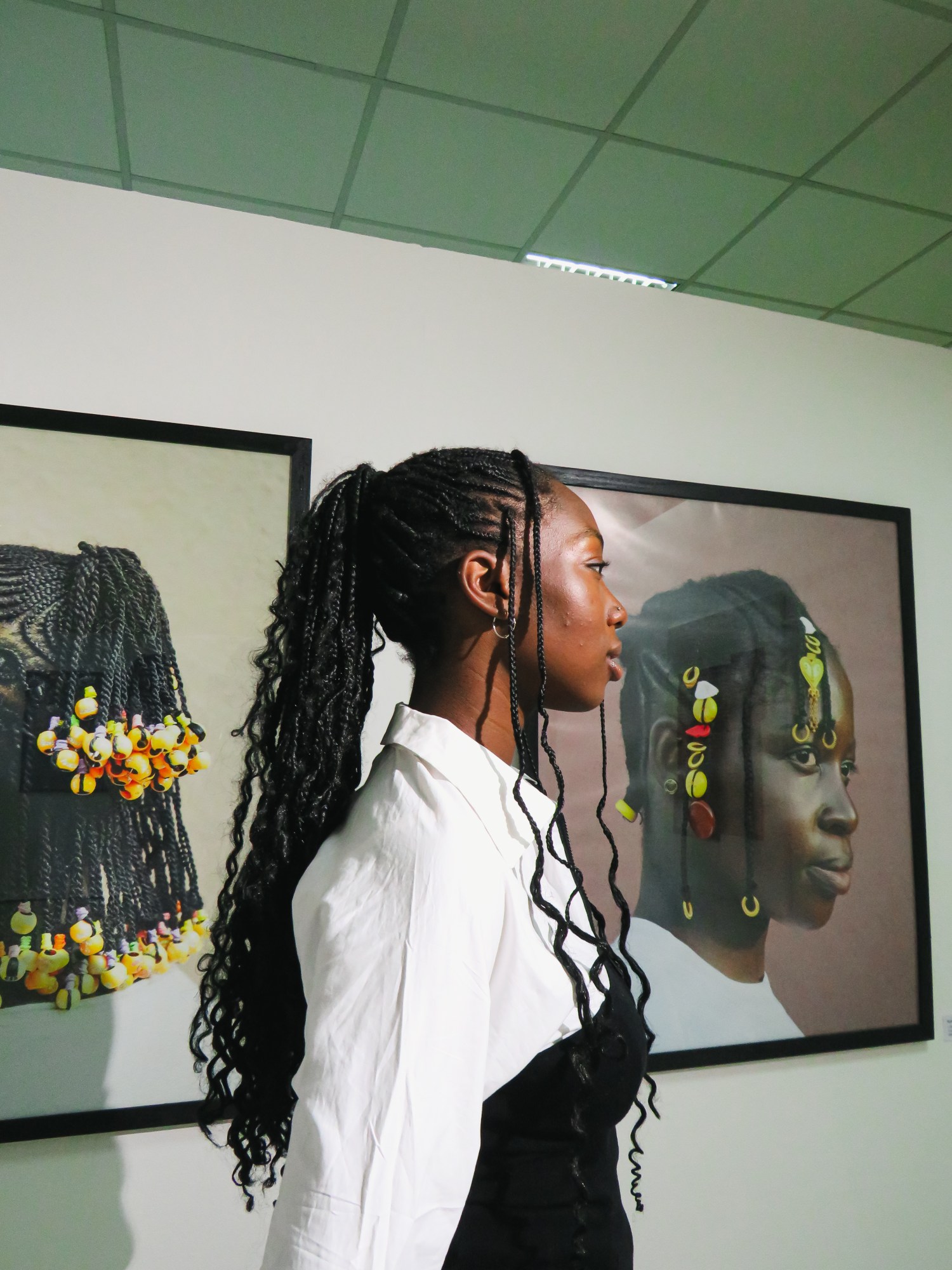 A young Black woman with long braids stands side-on in front of two exhibition portraits of women with similar braids.