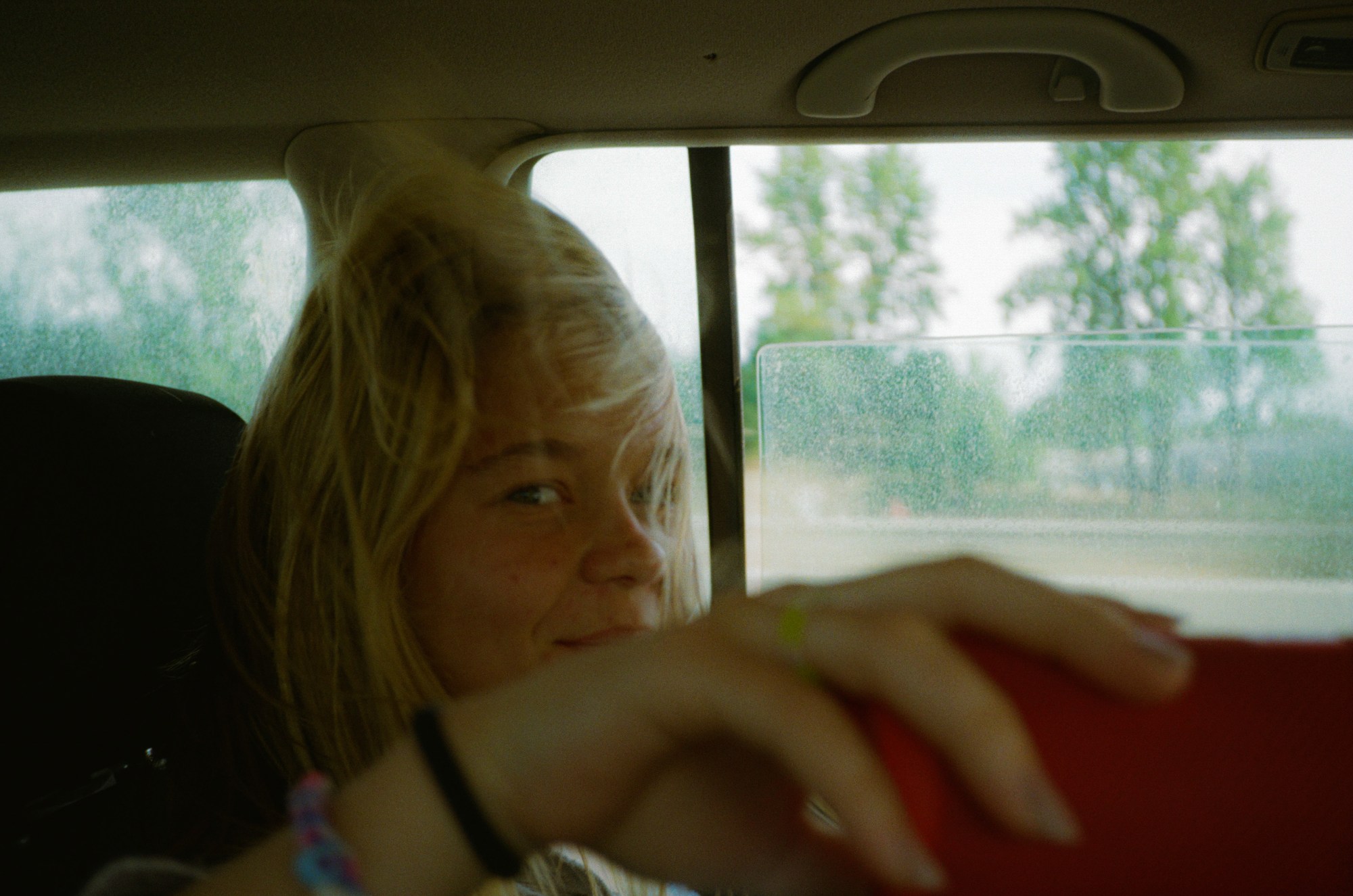 A young person with blonde hair sits in the backseat of a car; the window is down and her hair is blowing across her face. She smiles slightly .