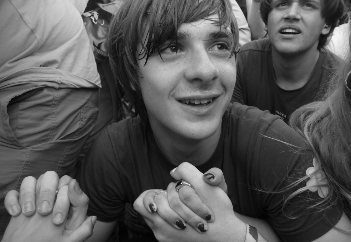 a black and white image of a smiling person with cropped hair holding hands with two unseen friends in a mosh pit; he is surrounded by other people