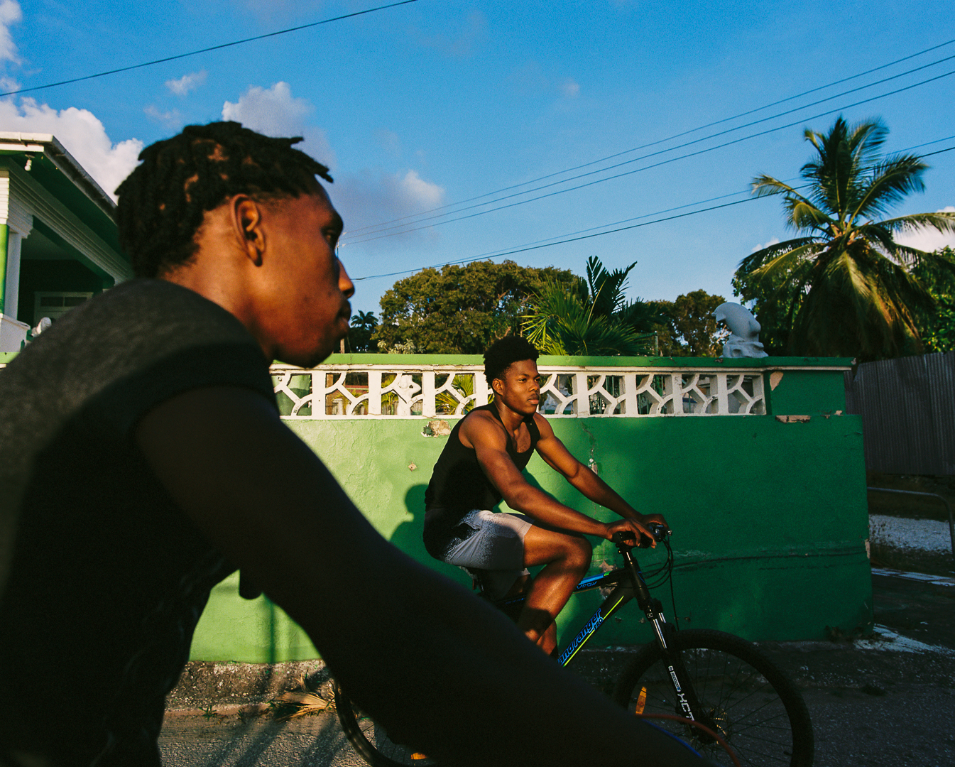 Two young Black men ride bikes along a sunny street in Moontown, Barbados. Behind them is a green wall and a couple of palm trees set against a bright blue sky.