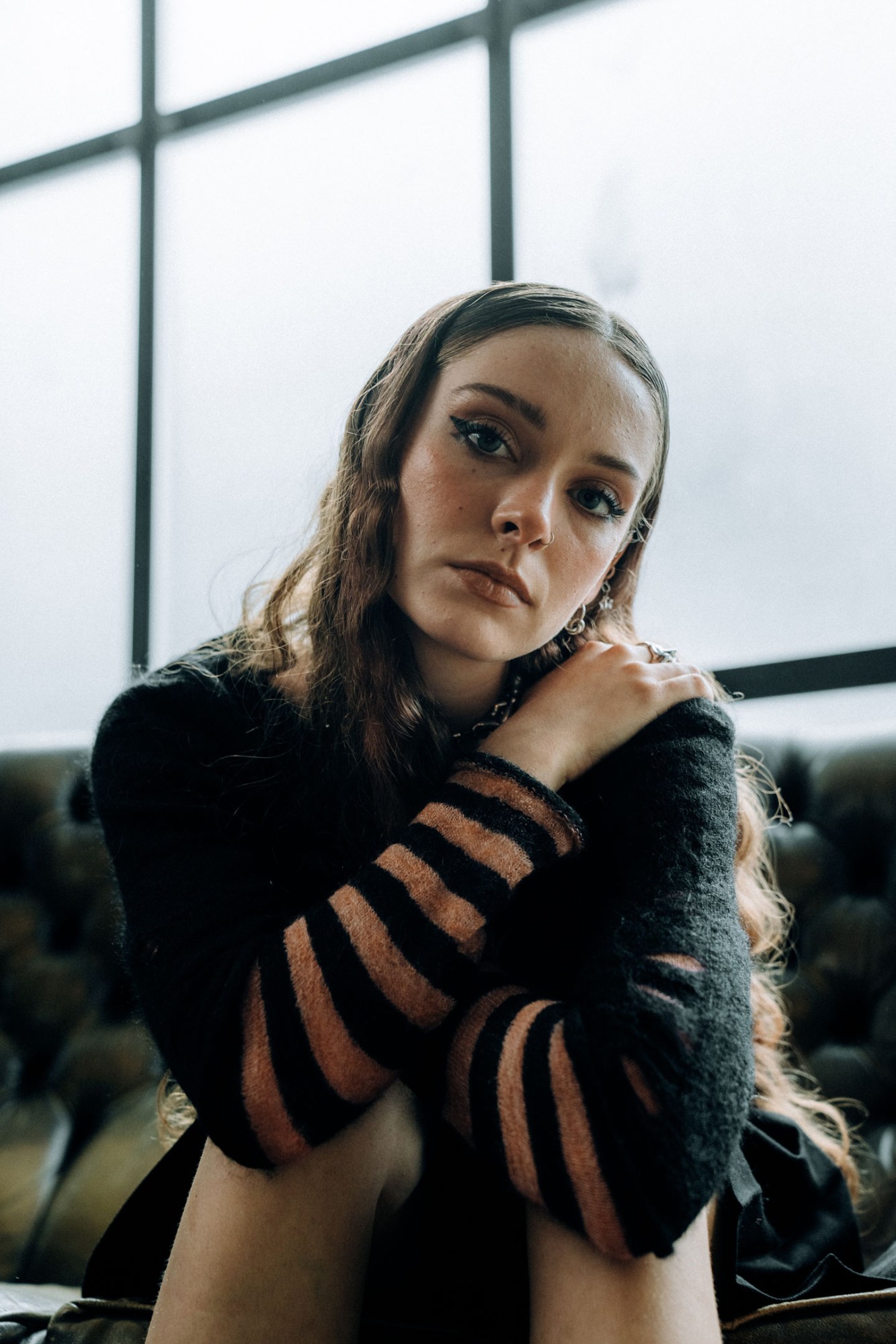 holly humberstone in a striped sweater, hugging her legs to her chest on a sofa and looking into the camera