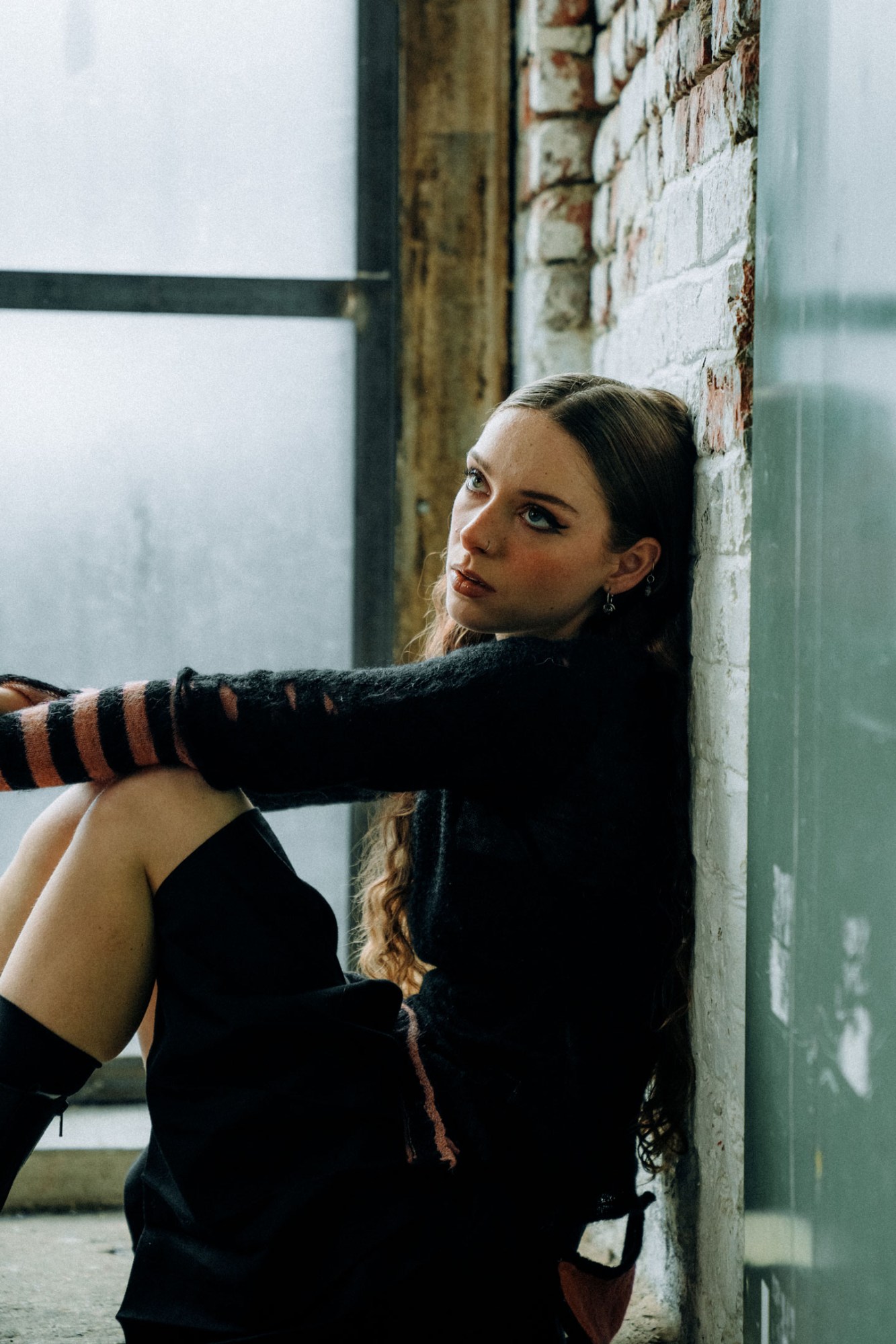 holly humberstone sitting side-on against a tiled wall, she's wearing a skirt and striped sweater and is looking off camera with an annoyed expression