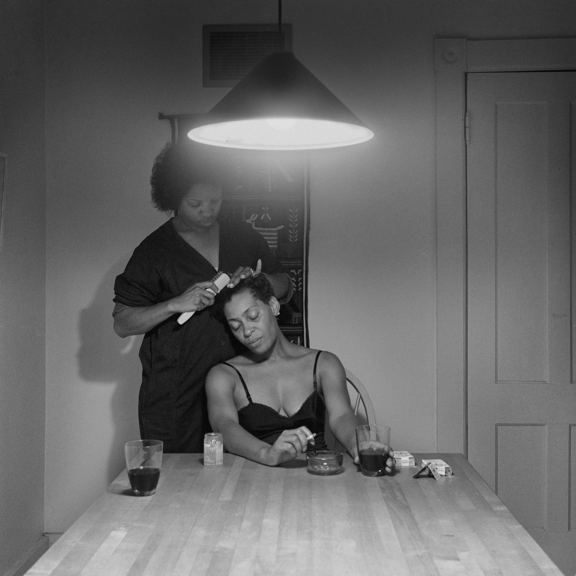 Carrie Mae Weems sitting at a kitchen table drinking wine with a friend who is brushing her hair