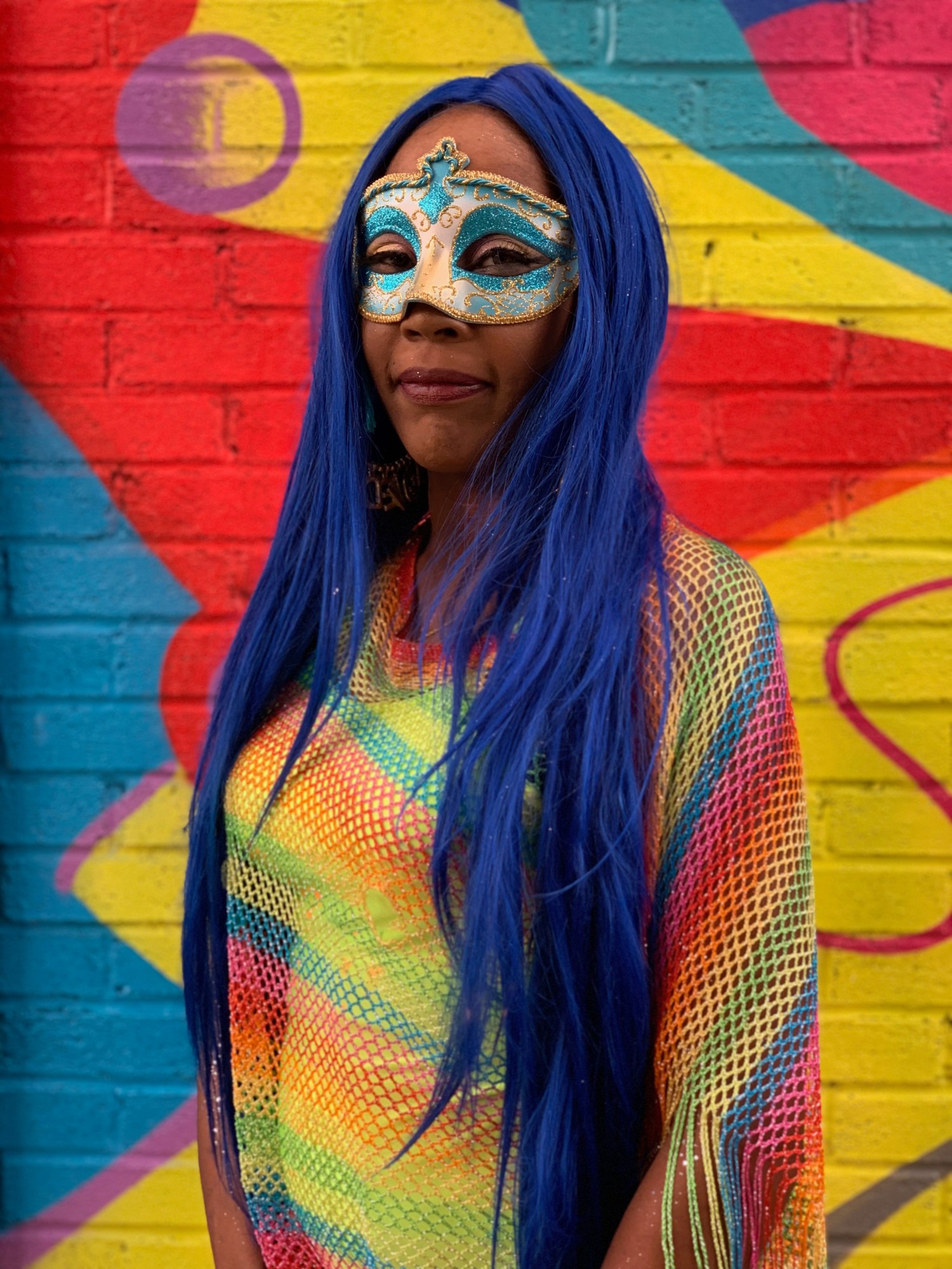 Photograph from End of the Line by Taylor Chapman of a woman with blue hair in a rainbow net top and a mask against a painted wall