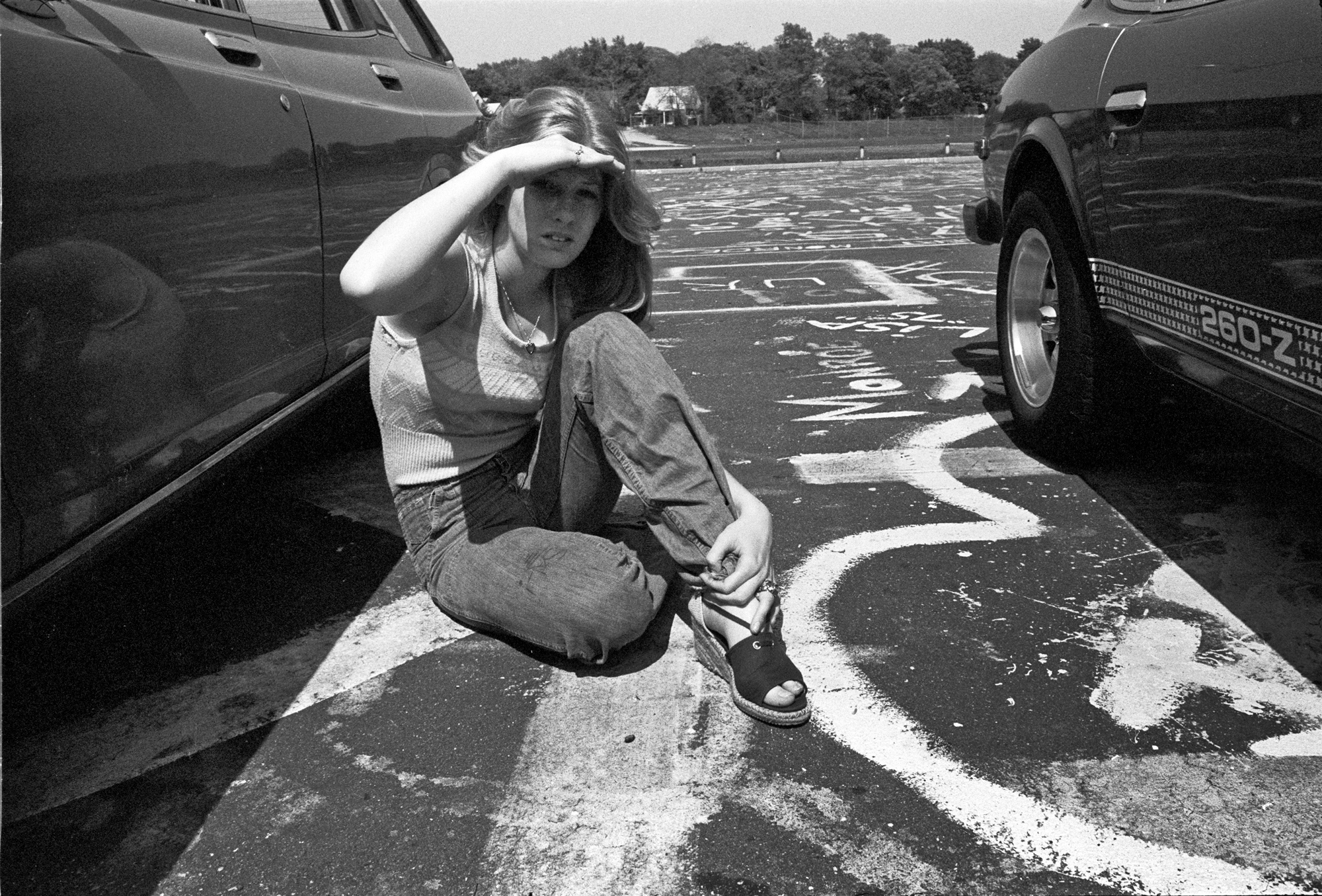 a girl sitting down between two cars in the high school parking lot in 1976