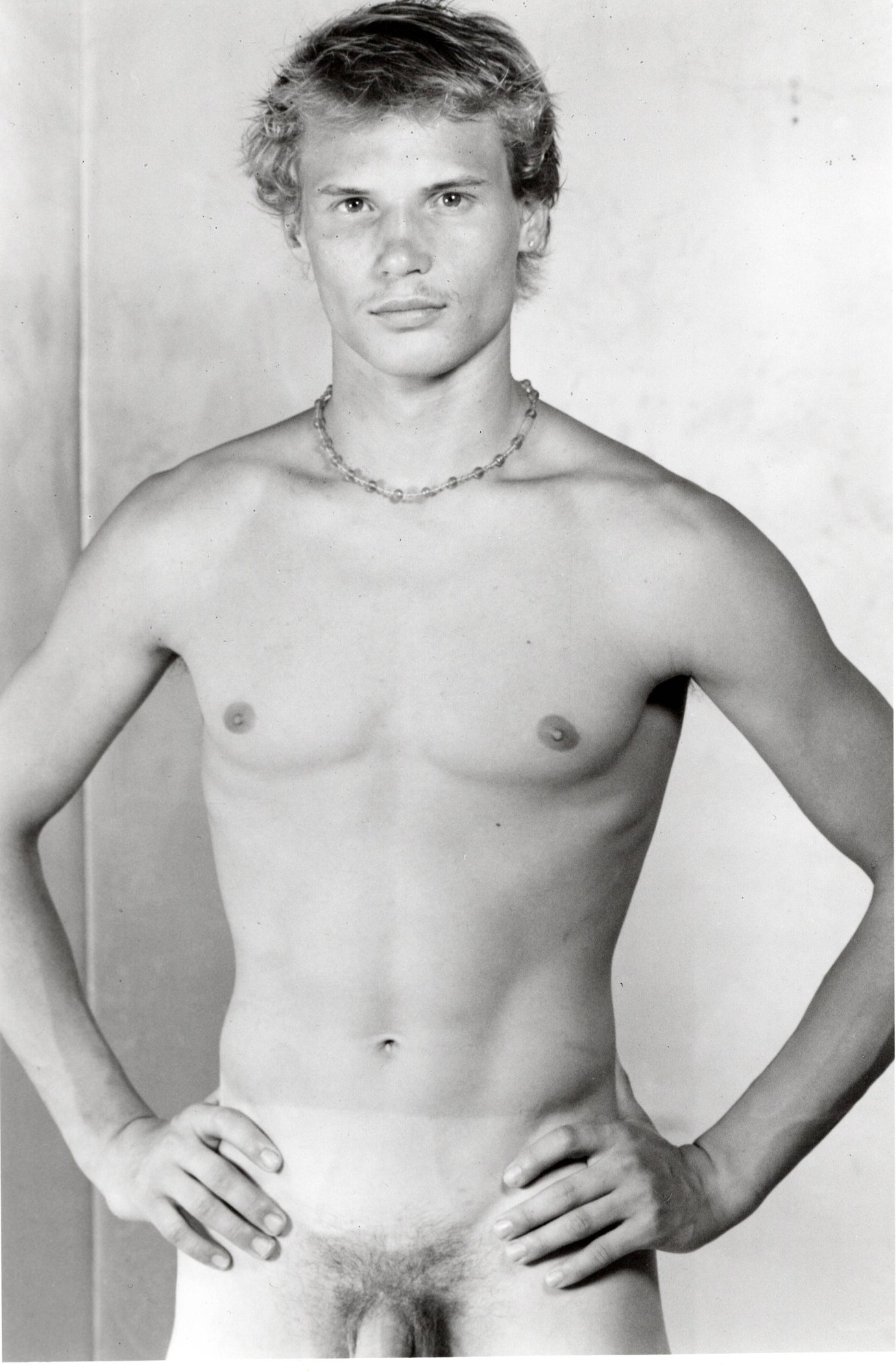 a full frontal b+w portait of a nude man with his hands on his hips. he is young-ish, with surfer hair and a beaded necklace