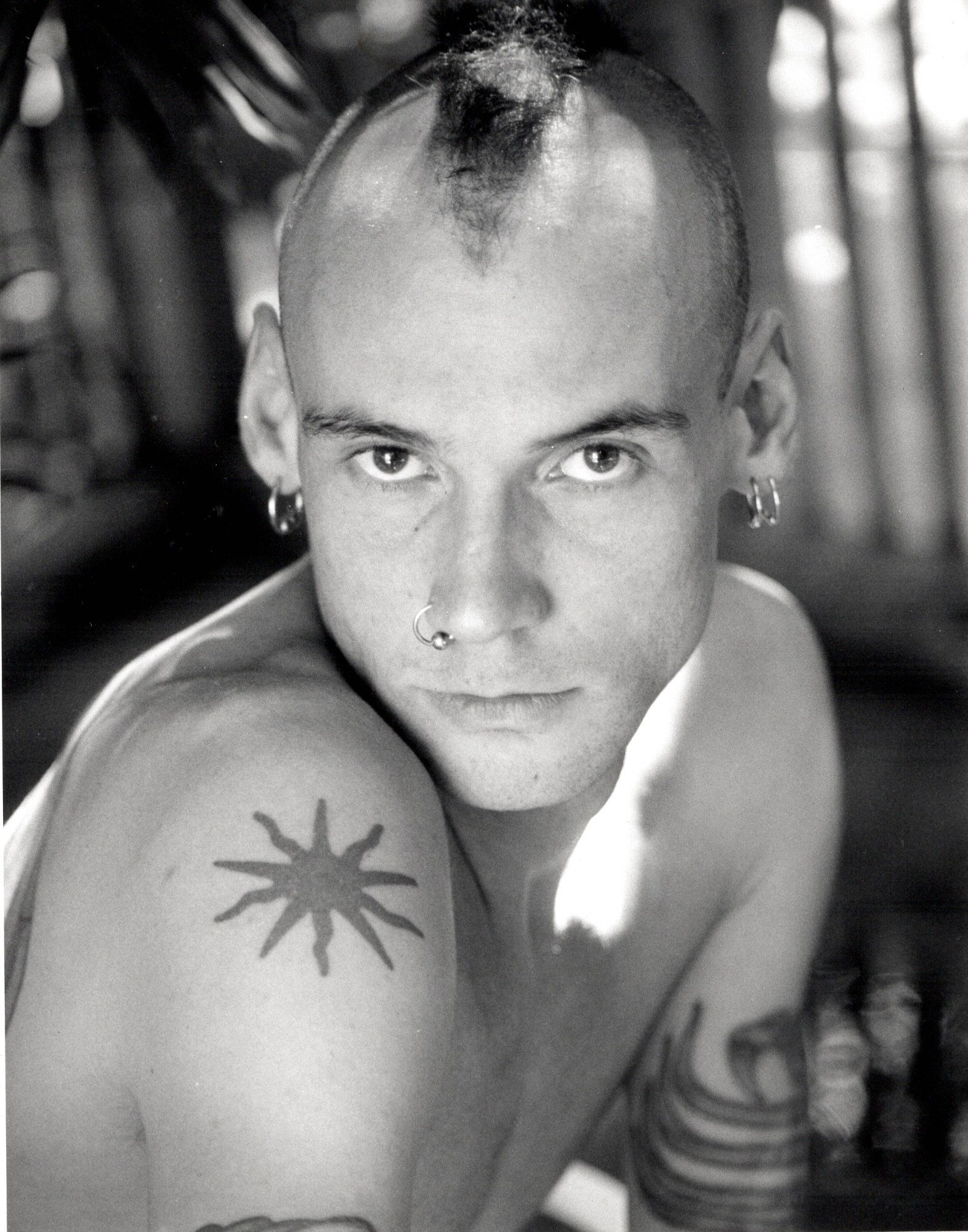 a man with a mohawk, multiple piercings and a tattoo of a sun on his upper arm. he is topless and looks to the camera seductively