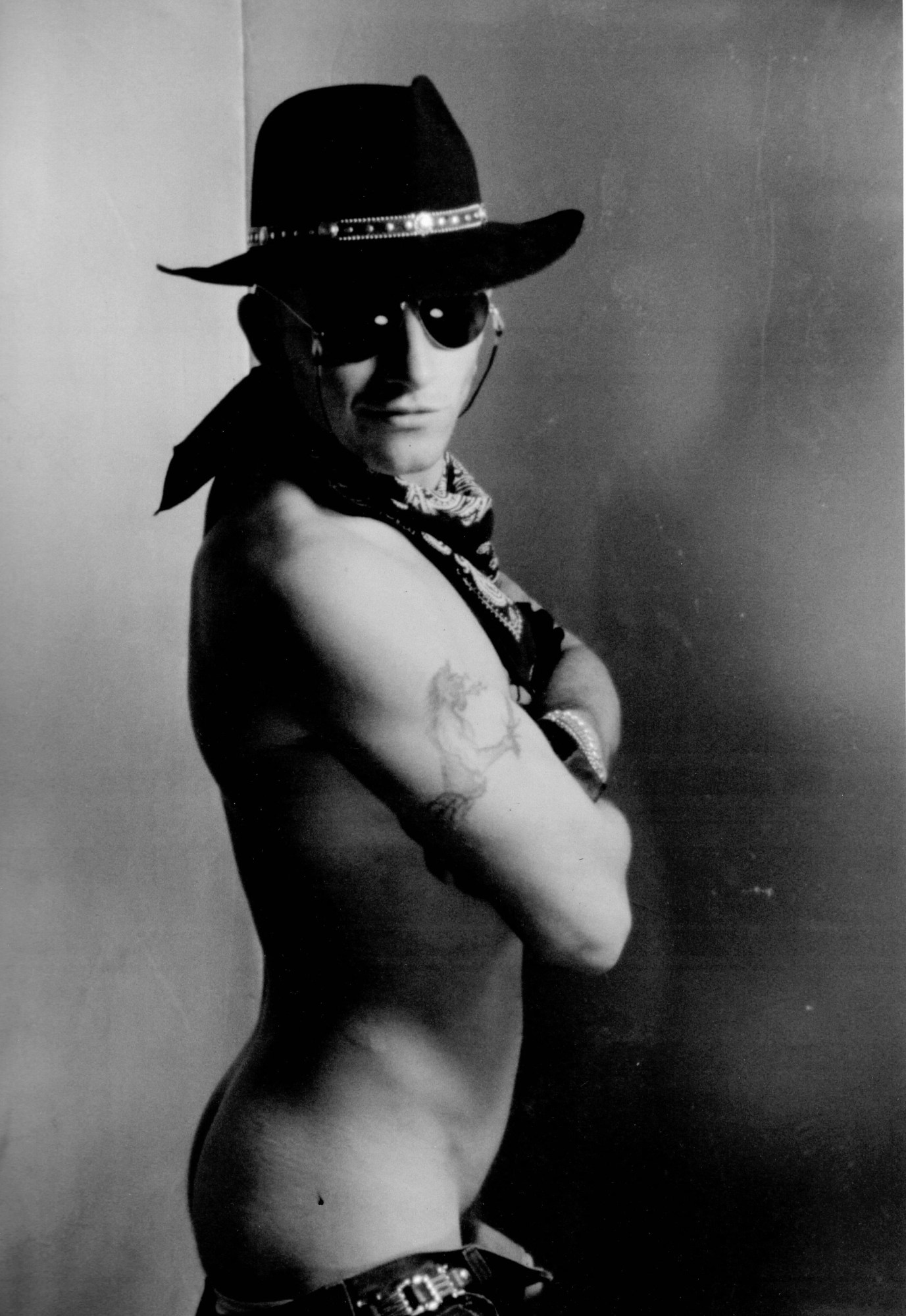 a nude man wearing thigh-high boots (just visible) and a cowboy hat stands side-on with his arms crossed, posing in a photo studio