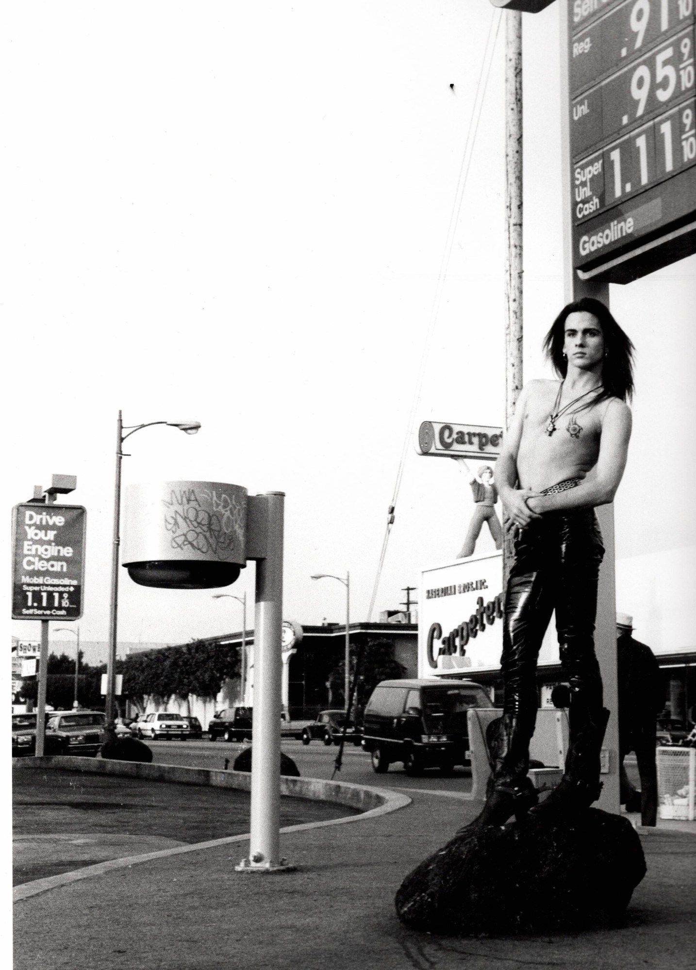 a topless man with long hair and tight leather trousers stands on a rock on the side of an LA street, signs and advertisements clutter behind him