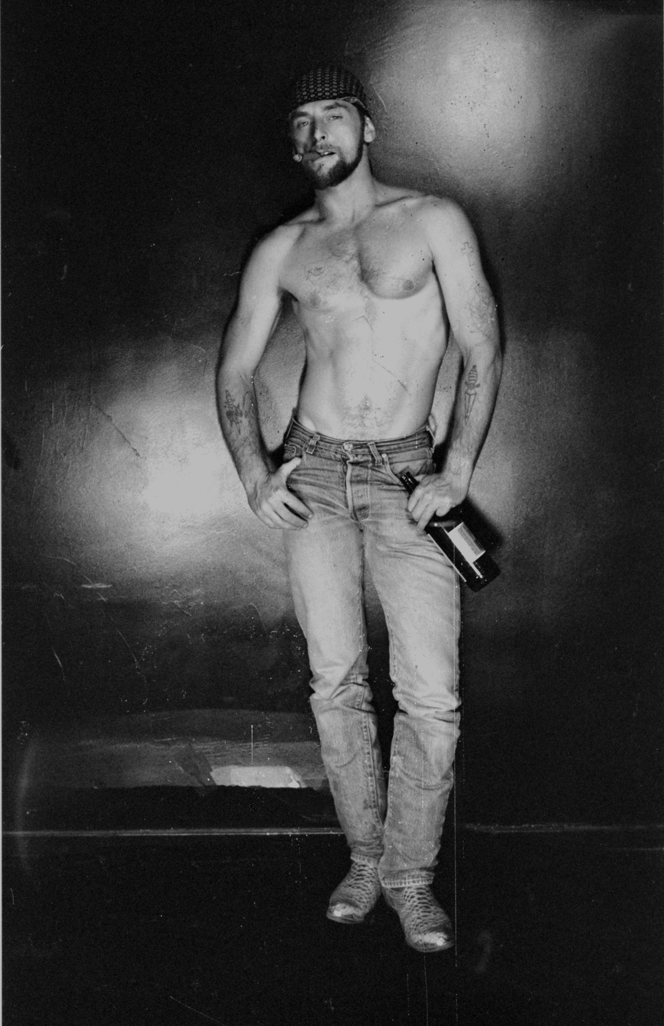 a topless bearded man in a pair of jeans holds of bottle of beer in one hand and a cigar between his teeth