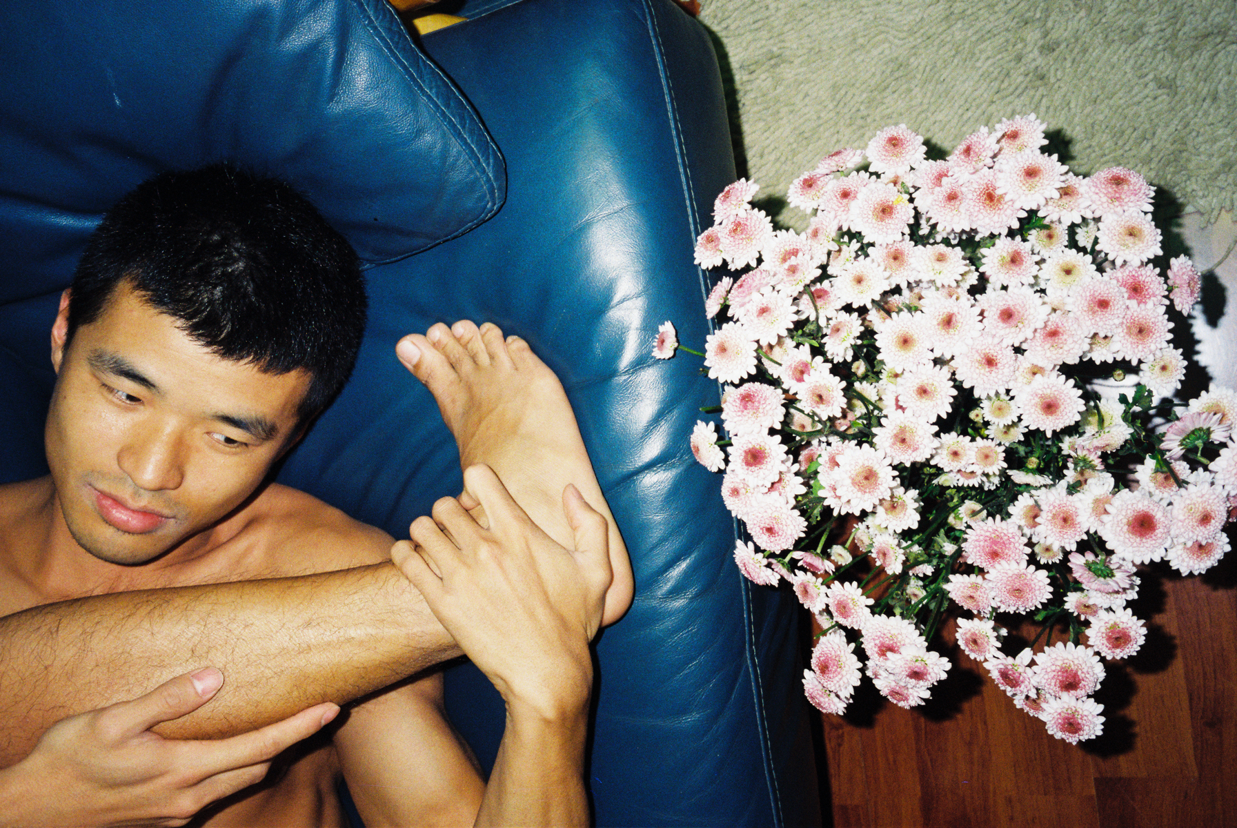 lin zhipeng photograph of a man with his leg stretched over his body, up to his neck. he's naked. a bunch of flowers sit on a table next to him.