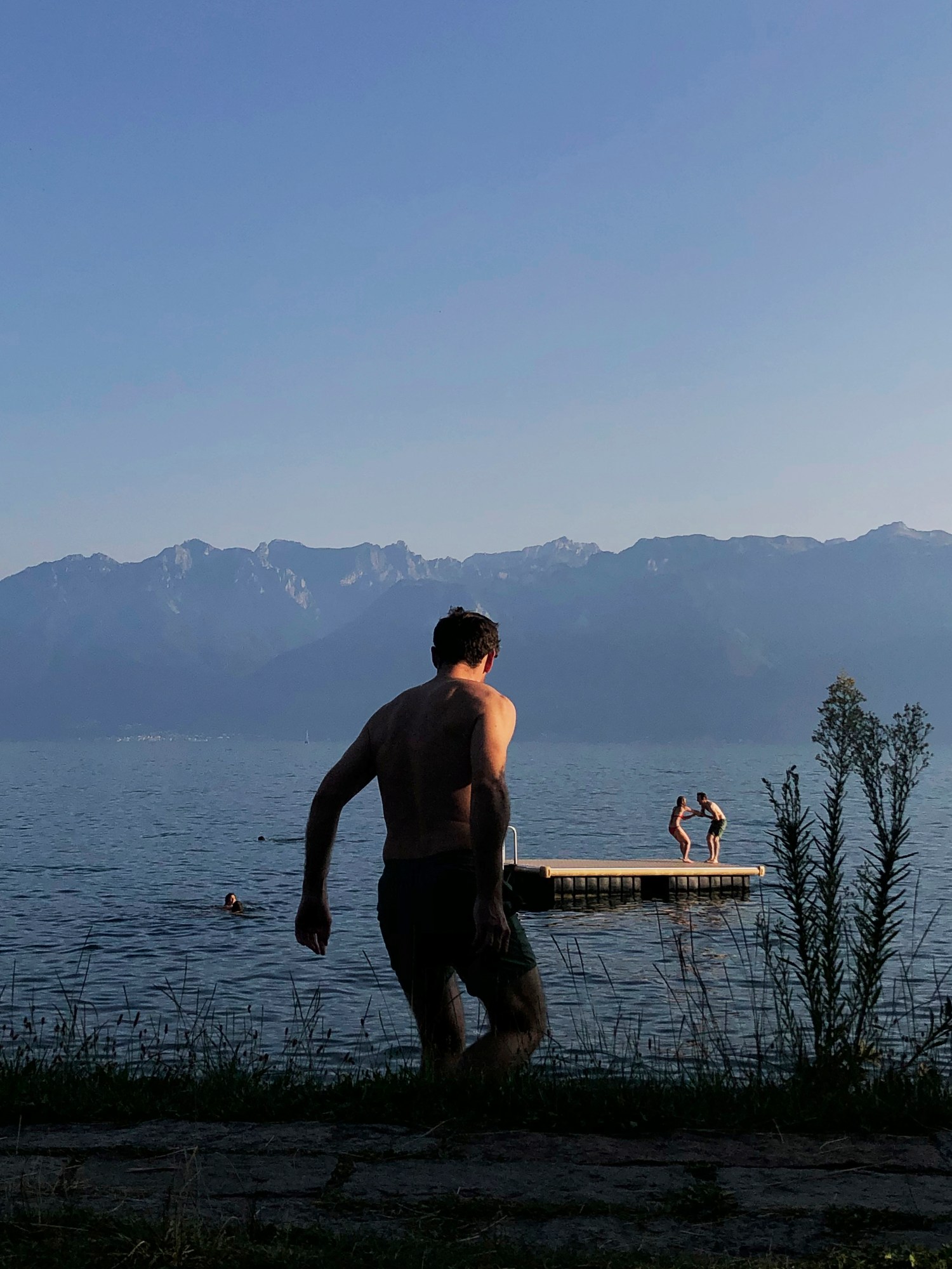 A man seen from behind walking towards a lake at sunset, two people in the background play on a jetty