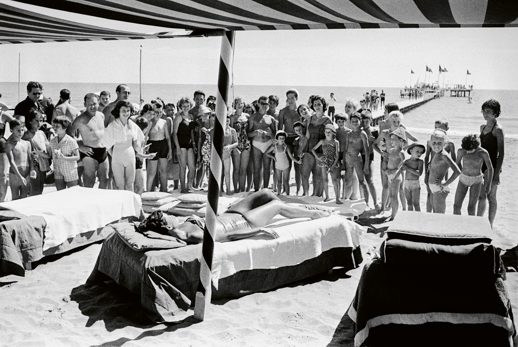 a black and white photograph by paolo di paolo of a woman on the lido beach at venice film festival. she sunbathes as a crowd watch on.