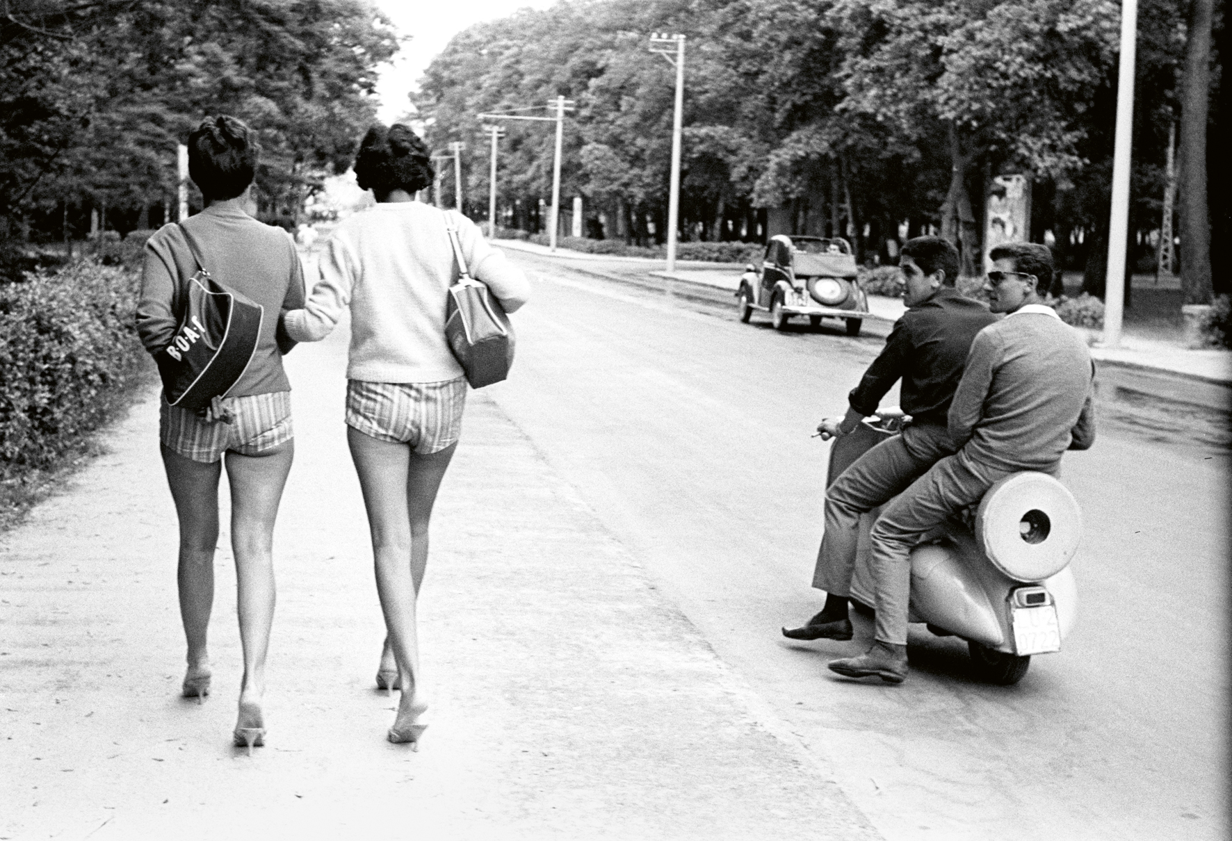 a black and white photograph by paolo di paolo of two women walking down the street in shorts, sweaters with bags on their shoulder. two men on a moped watch on.