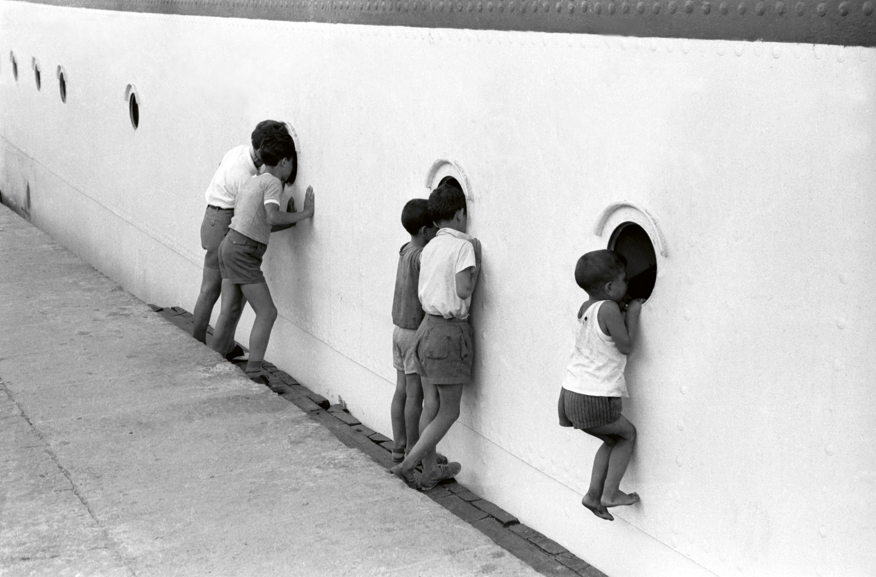 a black and white photograph by paolo di paolo of children on port of messina peering through the holes of a boat.