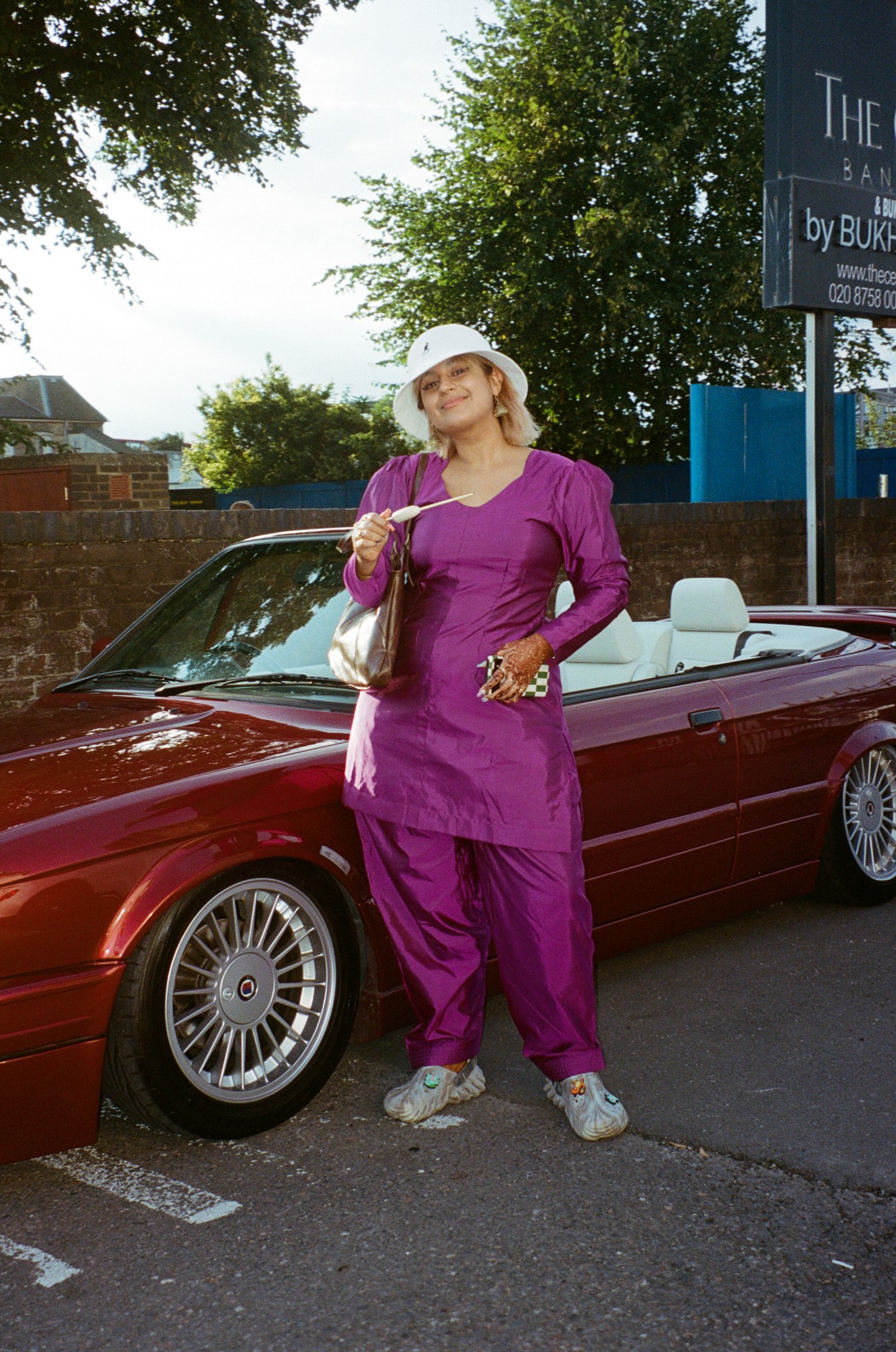 a young south asian woman in a matching magenta outfit wears a white bucket hat and poses holding a kulfi in front of a classic red BMW car