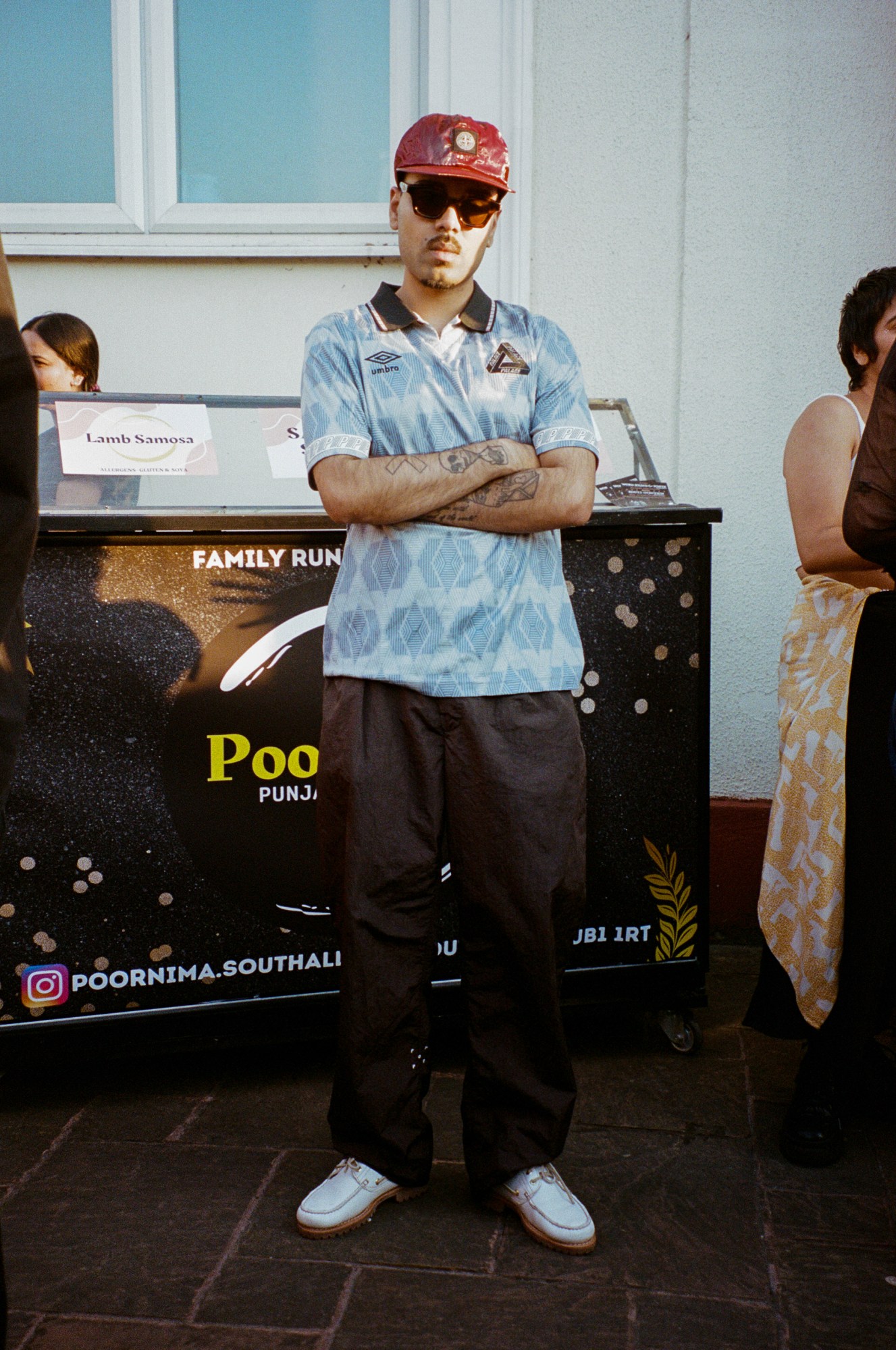 a young south asian man in a palace x umbro baby blue football shirt crosses his arms and looks to camera