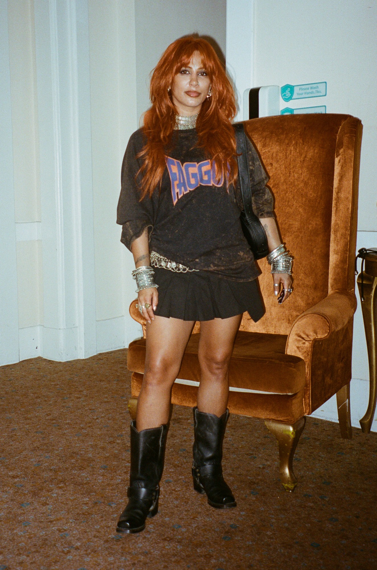 a south asian woman with orange hair wears cowboy boots and a jumper that says faggot