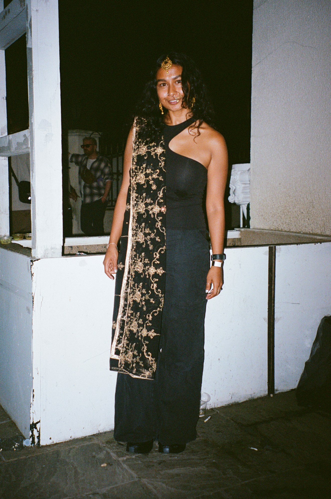 a young woman in a long black dress with a black and gold scarf over one shoulder smiles to camera