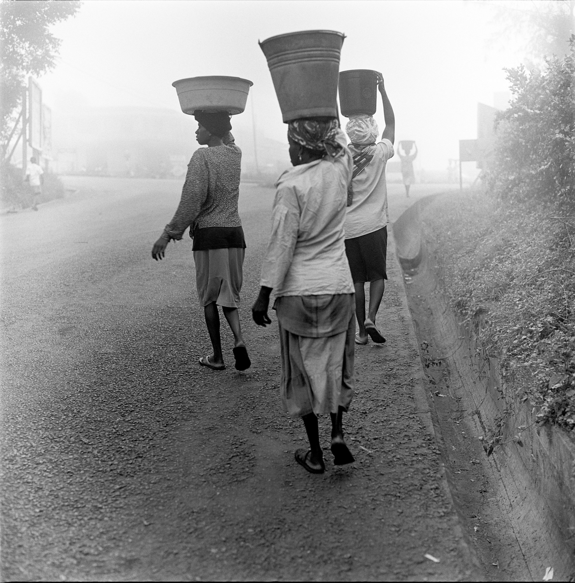 black-and-white photo of three women carrying buckets on their heads walking away from the camera