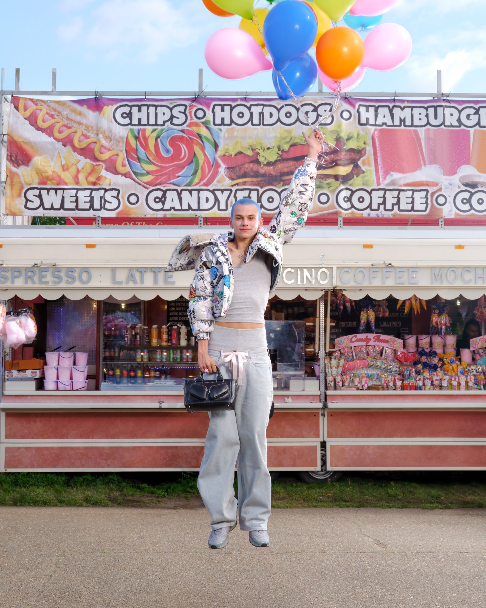 a model with a bright blue buzzcut and a bomber jacket appears to float above the ground thanks to the bunch of balloons he holds above his head