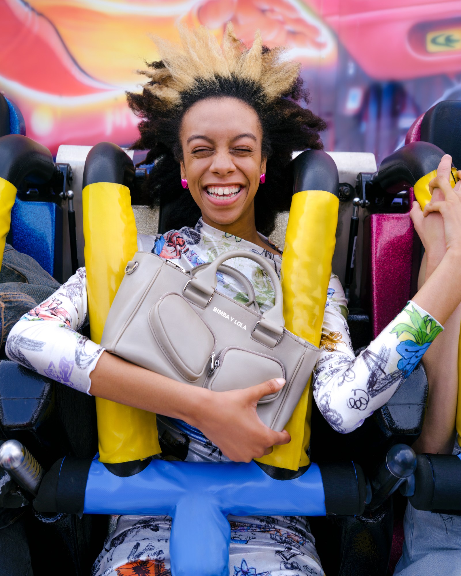 a model with pink heart earrings and a graffitied silk dress smiles as they clutch a taupe leather handbag on a fairground ride