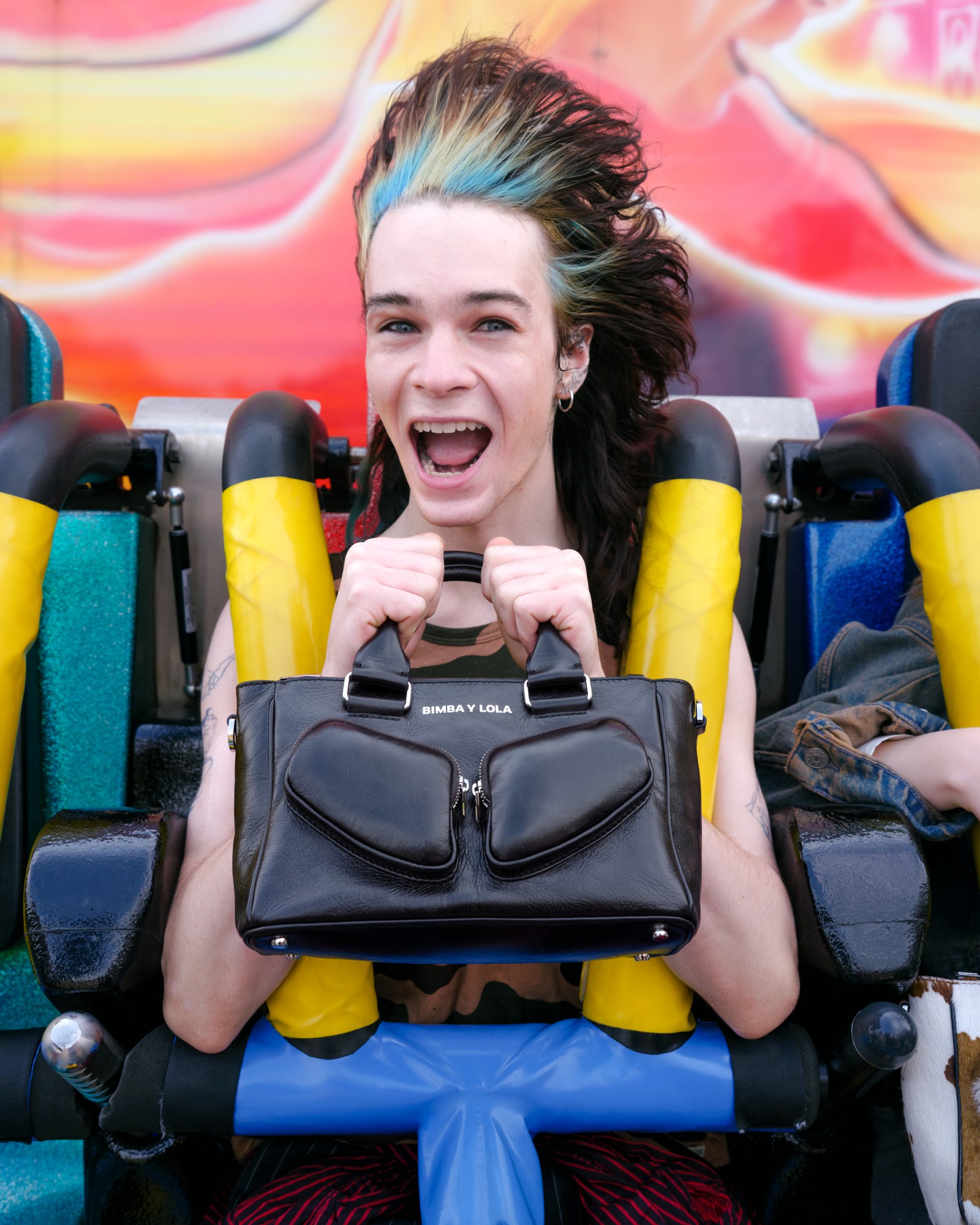 a model with blue and bleached hair streaks clutches a black leather handbag on a fairground ride