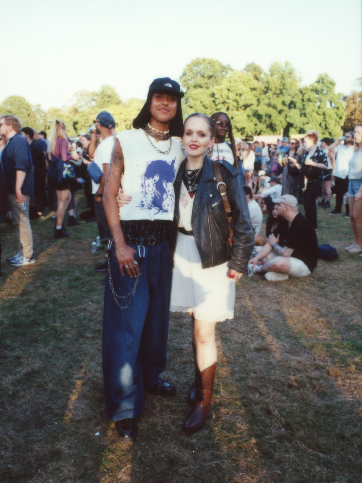 a couple with their arms around each other posing for a photo; one is wearing jeans with chains and a boygenius merch tee; the other a leather jacket over a dress photographed by Jody Evans